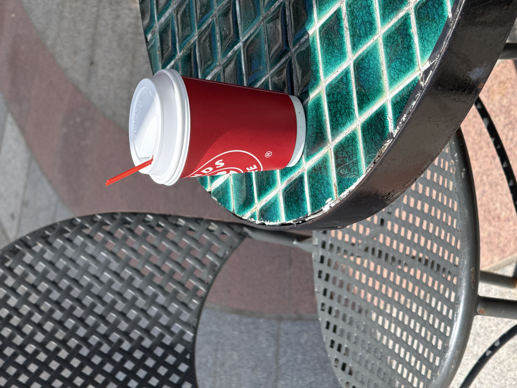 A red paper soda cup with a white lid and red straw rests on a small, round table with turquoise, diamond-shaped tiles and a black metal edge. The table sits between two metal chairs with a woven, gray pattern. The ground is made of concrete and red brick.