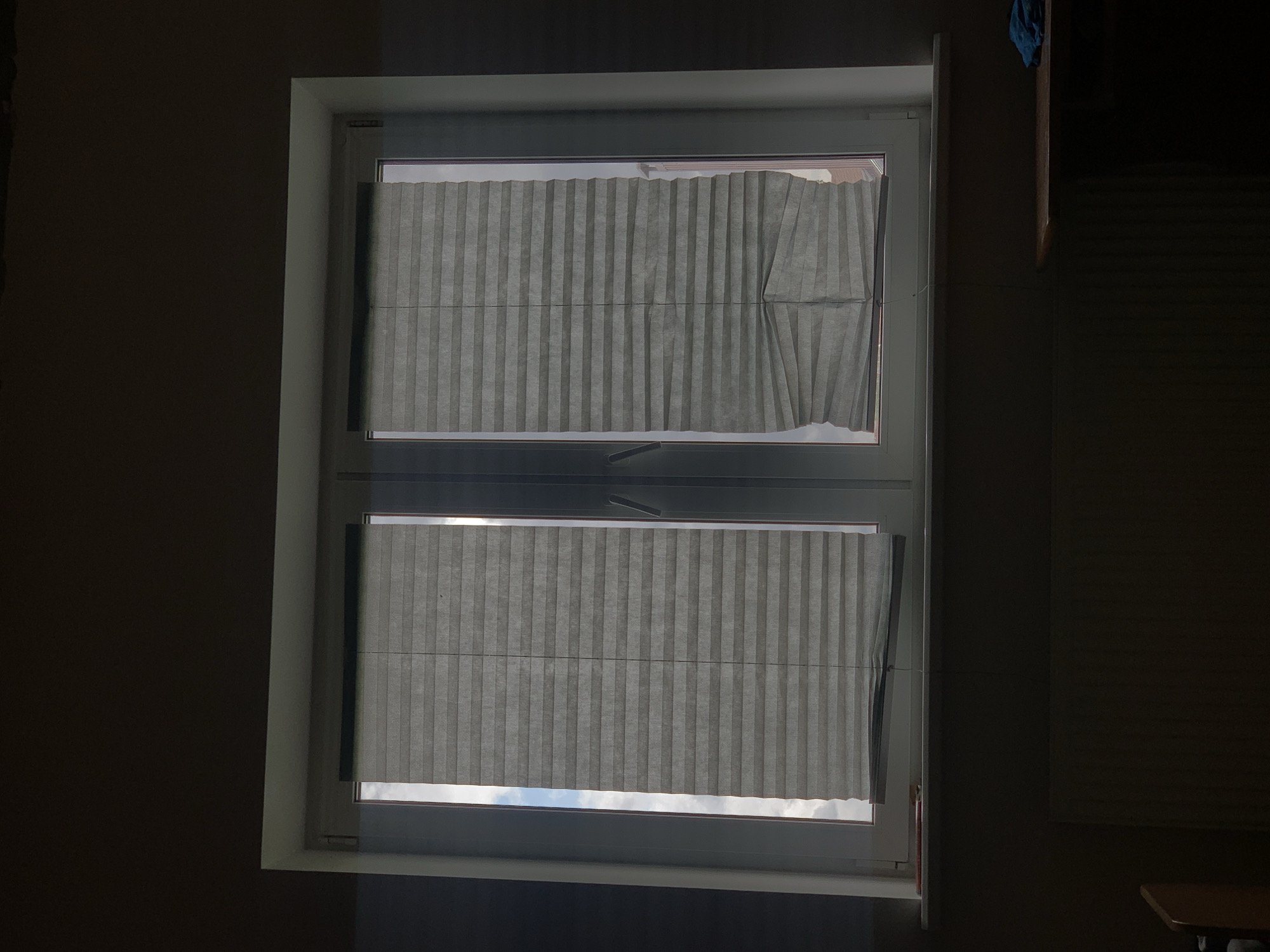 The image shows a window with grey pleated blinds. The blinds are partially drawn, covering the majority of the windowpane. The left blind appears to be fully drawn down, while the right blind is drawn down to about two-thirds of the way. The right blind also shows some distortion or damage at the bottom, with folds not aligning properly. Through the uncovered portion of the window, a cloudy sky can be seen, suggesting it is daytime. The window itself is framed by a white structure. Below the window, a radiator is visible, and to the right, a portion of a wooden shelf with a blue cloth is present. The surrounding walls are dark, indicating an indoor setting, likely a room. The scene is dimly lit, with light filtering in from the window. The image is an interior shot of a residential or office space. Without additional context, it is impossible to determine the precise location within Minsk, Belarus.
