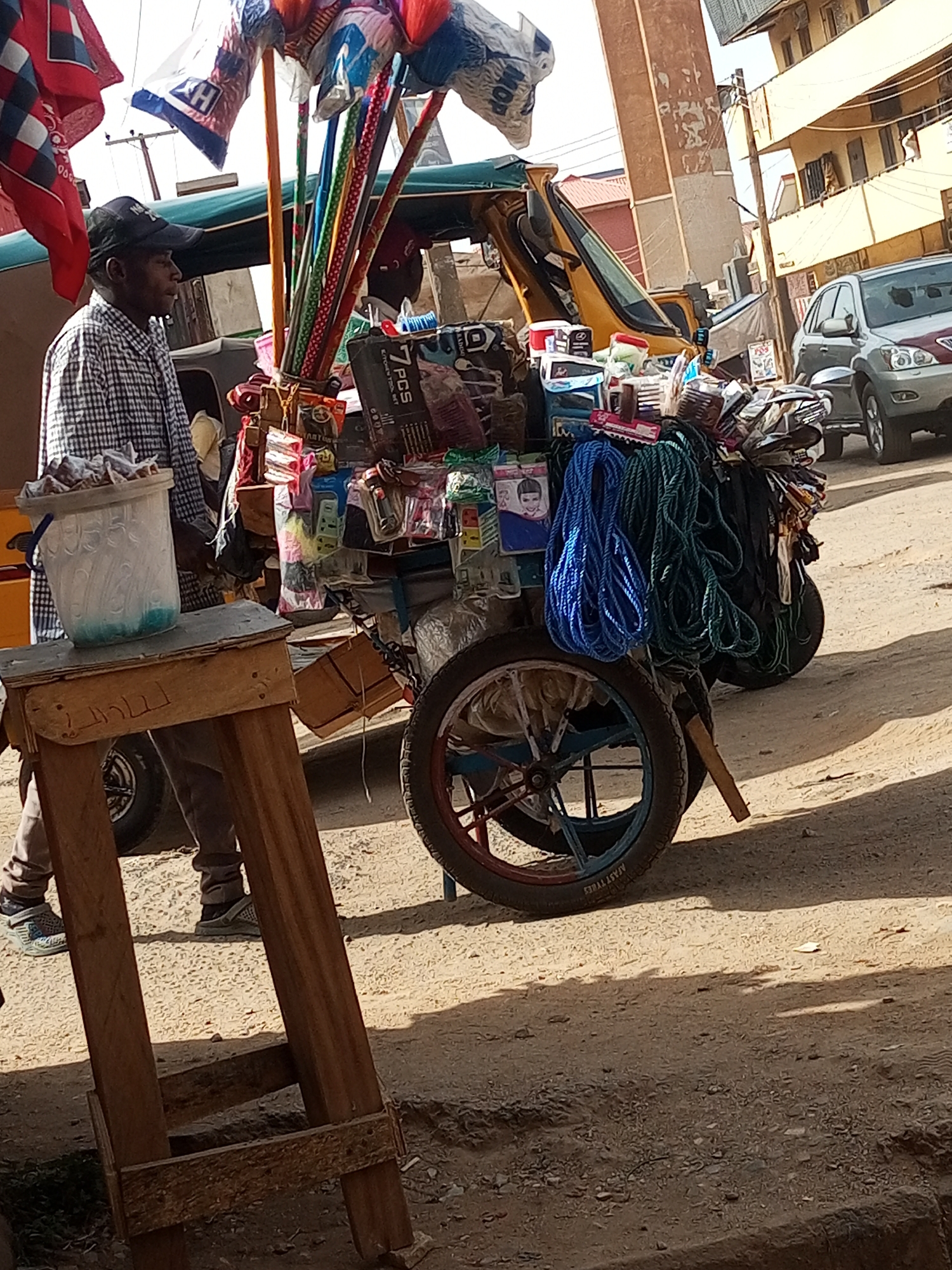 The image shows a street vendor in Maiburiji, Nigeria, standing next to his hand-pushed cart laden with goods for sale. The vendor, a Black man wearing a checkered shirt and a baseball cap, is looking to the left. The cart is heavily stocked with various items including mops, ropes, packaged goods, and tools. A small wooden stool with a plastic bucket on top is in the foreground. In the background, there are buildings and parked vehicles, including a yellow auto rickshaw and a gray SUV, indicating an urban setting. The ground is dirt and possibly asphalt. The lighting suggests it is daytime.