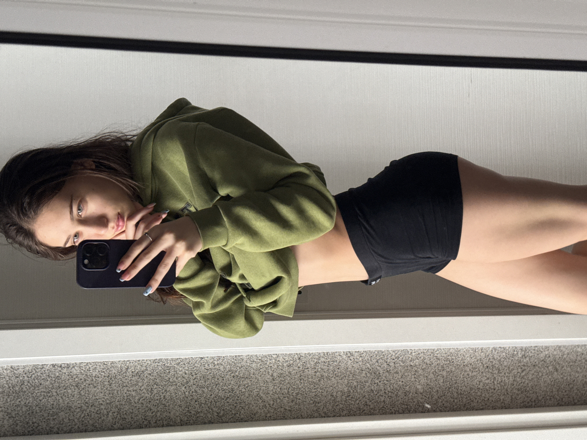 An indoor, full-body mirror selfie features a young woman rotated 90 degrees clockwise. She has long, dark hair and light-colored eyes. She is wearing a loose-fitting, olive-green hooded sweatshirt and black athletic shorts. A dark-colored smartphone, held in her left hand, covers part of her face, and she looks directly into its camera, suggesting she is taking the photo. Her right hand rests near her chin. A silver ring is visible on her right ring finger, and her fingernails are painted light blue, with some showing a subtle design.
The background, visible as a reflection, consists of a white wall or ceiling above a light-colored trim, and a grey, textured carpet or floor below. The lighting is bright and even, indicative of an indoor setting. There are no clear cues within the image to suggest the specific location of Sofia, Bulgaria. No legible text is visible on her clothing or the phone. The woman's expression is neutral to slightly pensive.