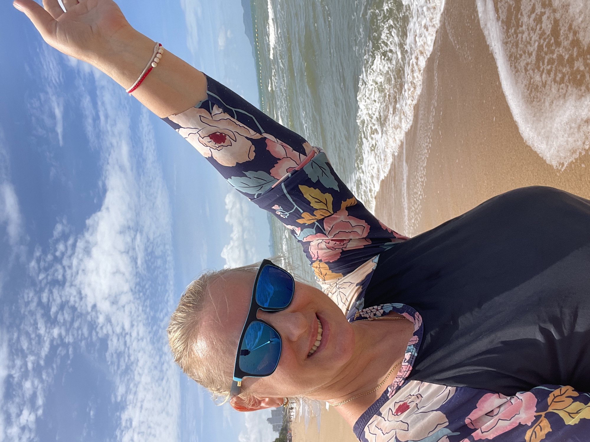 A Caucasian woman with blonde hair pulled back is taking a selfie on a beach in Pattaya City, Thailand. She is wearing sunglasses and a dark blue rash guard with a floral print on the sleeves. Her right arm is raised, and she is wearing a bracelet with beads. The background shows the ocean with waves and a cloudy blue sky. In the distance, there are buildings along the coastline. The sand on the beach is light brown.