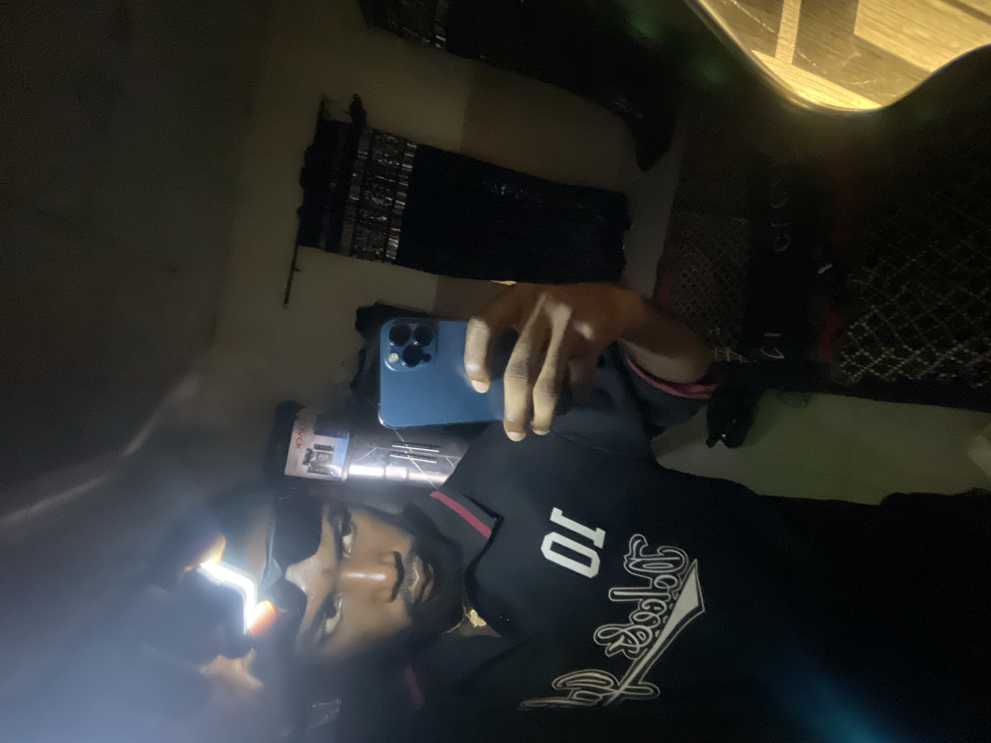 An indoor mirror selfie taken in Lokoja, Nigeria, depicts a dark-skinned male subject. The subject, a young adult with a beard, wears dark sunglasses pushed onto his forehead. A strong, bright light source, possibly a headlamp or phone flashlight, is positioned above his forehead, causing significant glare. He wears a black polo shirt with dark red stripes on the collar and cuffs. The shirt displays a white number "01" and a reversed white script logo on the chest. A gold-colored chain with a pendant is visible around his neck. The subject holds a blue smartphone, consistent with an iPhone, in his right hand, reflecting his image in the mirror. His expression is serious, with pursed lips and eyes directed slightly to his right.
The background, reflected in the mirror, shows a light-colored wall. To the subject's right (viewer's left), dark curtains hang from a rod. To his left (viewer's right), a reflective surface shows a reversed "VICK" label on a box. The lower portion of the reflection indicates a patterned floor or rug and dark, possibly furniture, elements. The overall lighting is dim, augmented by the bright light source near the subject's head and the phone screen.