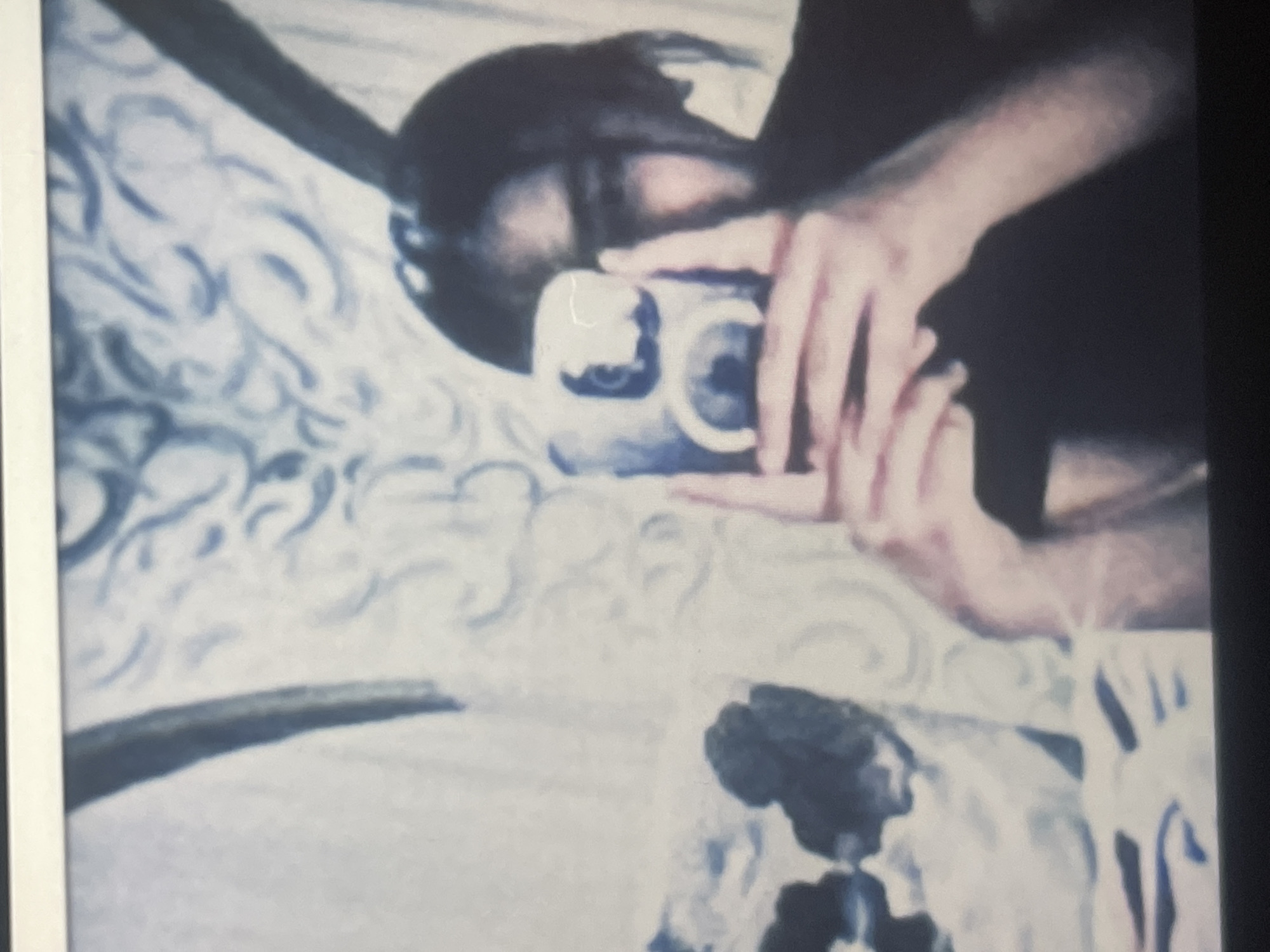This grainy, low-resolution image, possibly a photograph of an older print or screen, captures a person taking what appears to be a mirror selfie indoors. The main subject is partially visible, showing dark hair, a portion of their face, and both hands holding a light-colored camera or smartphone. The device is pointed towards the viewer, indicating a self-portrait being captured through a reflection.
The background features a wall or similar surface adorned with an ornate, swirling pattern in shades of light blue or grey against a white or very light background. The image is rotated, causing the pattern and subject to appear at an unusual angle. In the lower right corner, a dark, indistinct shape is present, possibly another element of the room's decor or an object.
The activity taking place is the act of photography, specifically a self-portrait. Due to the blurriness and the camera obscuring most of the face, emotions are not discernible. No visible text is present in the image. The indoor setting in Poltava, Ukraine, is a key location cue, although the visual content itself doesn't offer specific geographical markers beyond generic interior decoration. The overall impression is one of a vintage or retro aesthetic due to its muted color palette and soft focus.