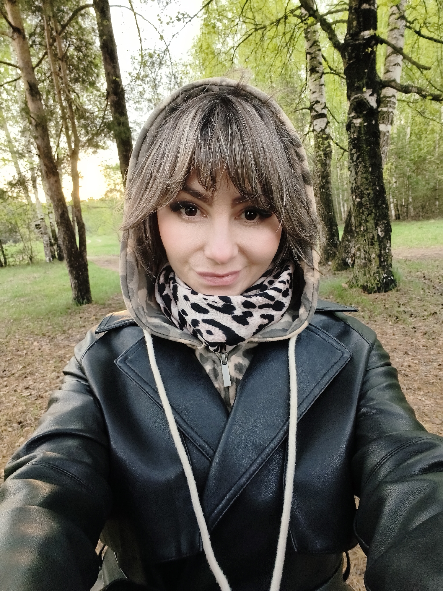 A woman is taking a selfie outdoors. She is wearing a black leather jacket, a camouflage hoodie, and an animal print scarf. The background consists of trees and grass, suggesting a park or forest. The location is likely in Chaikovsky, Russia.