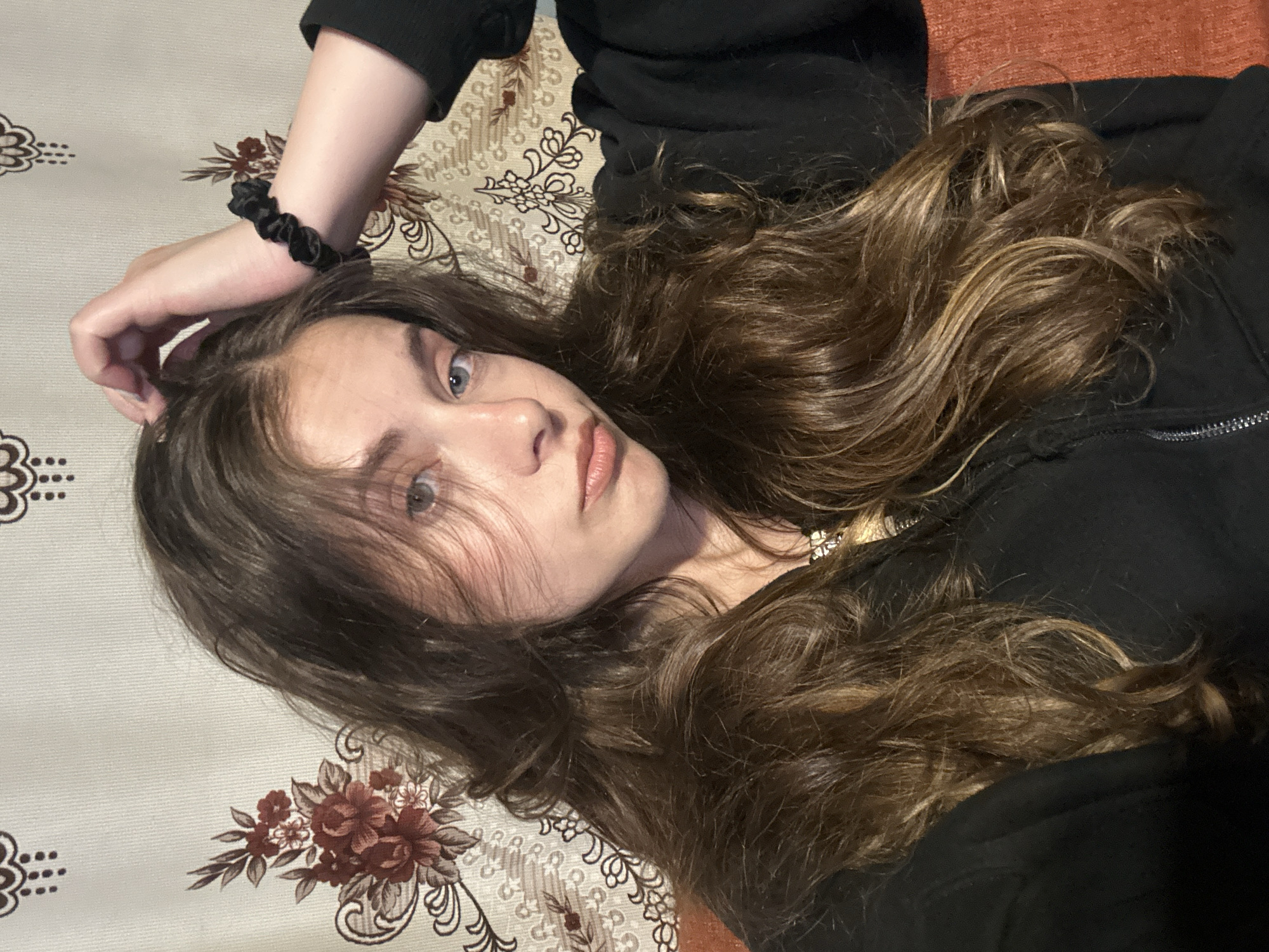 In the city of Unknown, Unknown, an indoor portrait captures a young female individual from the chest up, positioned slightly off-center and gazing directly at the camera. The subject has long, wavy, dark brown hair with lighter brown highlights, cascading over her shoulders and chest. Her eyes are blue/grey, and her lips are slightly parted in a pout. She wears a black hooded garment, partially revealing a silver zipper on the left side of her chest. Her left hand is raised, with fingers touching the top of her head near the hairline, and a black scrunchie is worn on her left wrist. A faint metallic necklace is visible at her collarbone. The background consists of a light beige or off-white wall covering featuring a repeating pattern of stylized floral motifs in dark red and brown, along with intricate scrollwork. A portion of an orange-brown object, possibly furniture or a cushion, is visible on the lower right side of the frame.