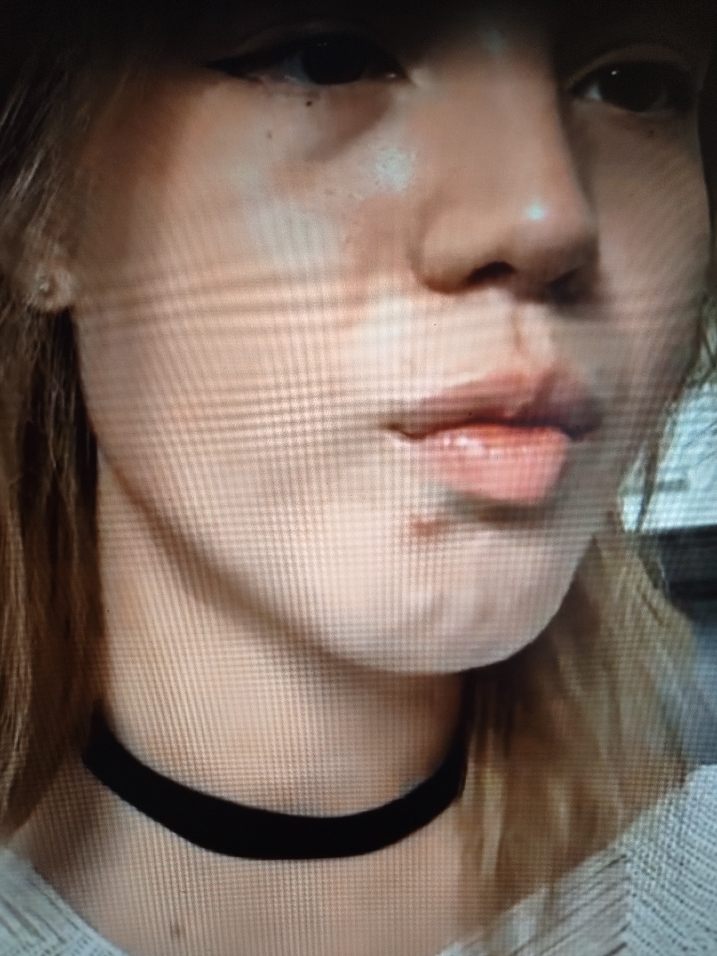 This is a close-up, eye-level, medium shot of a young person's face, focusing on the lower half of their face including their mouth and neck, and a portion of their right eye. The individual has fair skin with some subtle blemishes visible. They are wearing a black choker necklace. Their lips are plump and painted a light pinkish-red color. The background is dark and out of focus, with some lighter strands of blonde hair visible on the right side. The lighting is soft, highlighting the contours of the face. The image appears to be a still from a video taken in Il’inskiy, Russia, with the focus on the individual's facial features.
