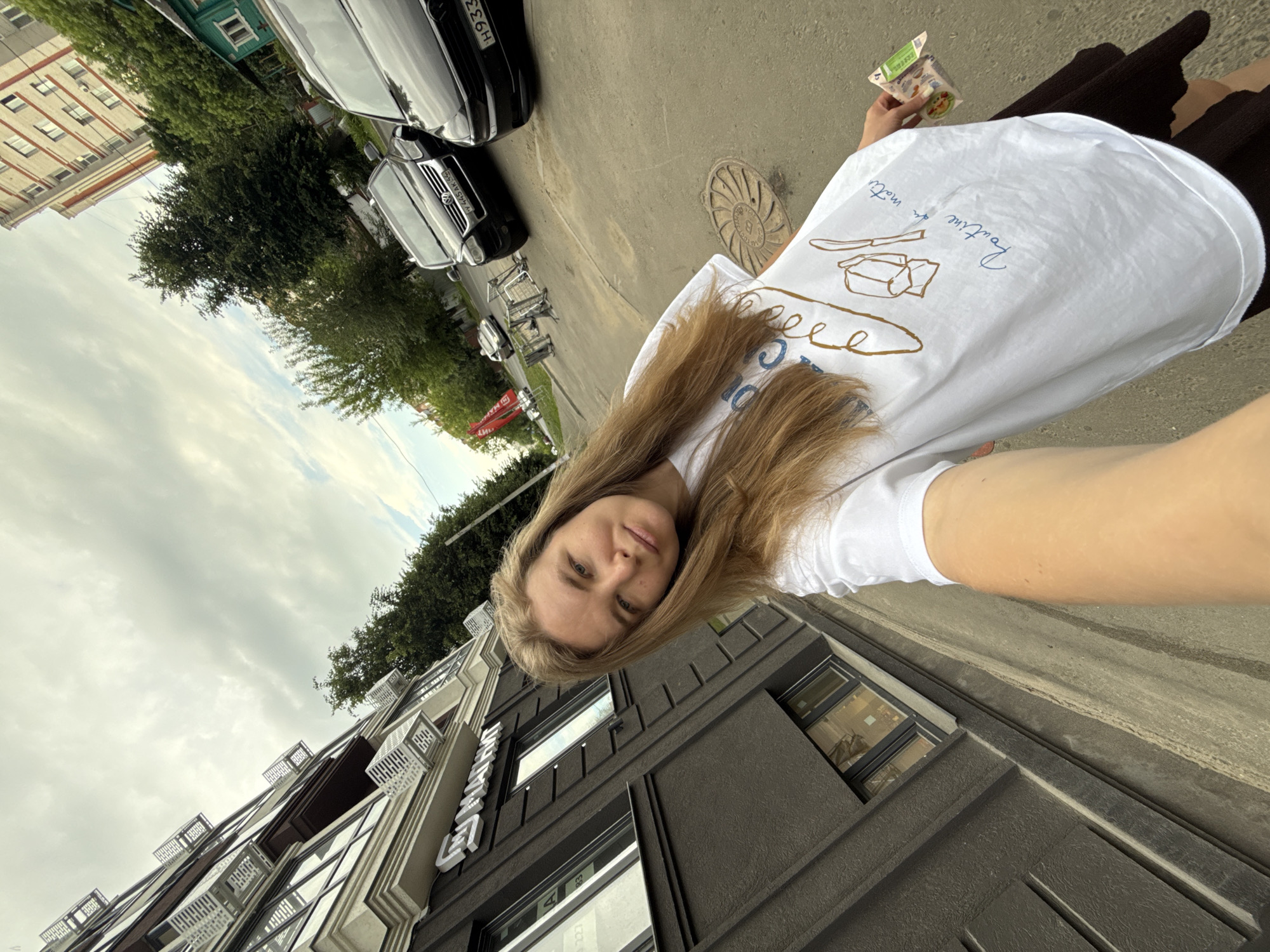 A young woman with long blonde hair is taking a selfie outdoors on a cloudy day. She is wearing a white t-shirt with a drawing of a spoon and fork and some blue text that says "Routine de matin" on it, and a dark skirt. She is holding a small package of what appears to be food in her right hand.
In the background, there is a modern building with dark grey and white facades, featuring multiple windows and balconies. To the right of this building, there are trees and a multi-story residential building with red brick accents. Several cars are parked on the street, including a black Volkswagen Touareg with a license plate that reads "VY 445 AK 55". A manhole cover is visible on the asphalt ground. The overall setting suggests a residential area within the city of Vladimir, Russia.