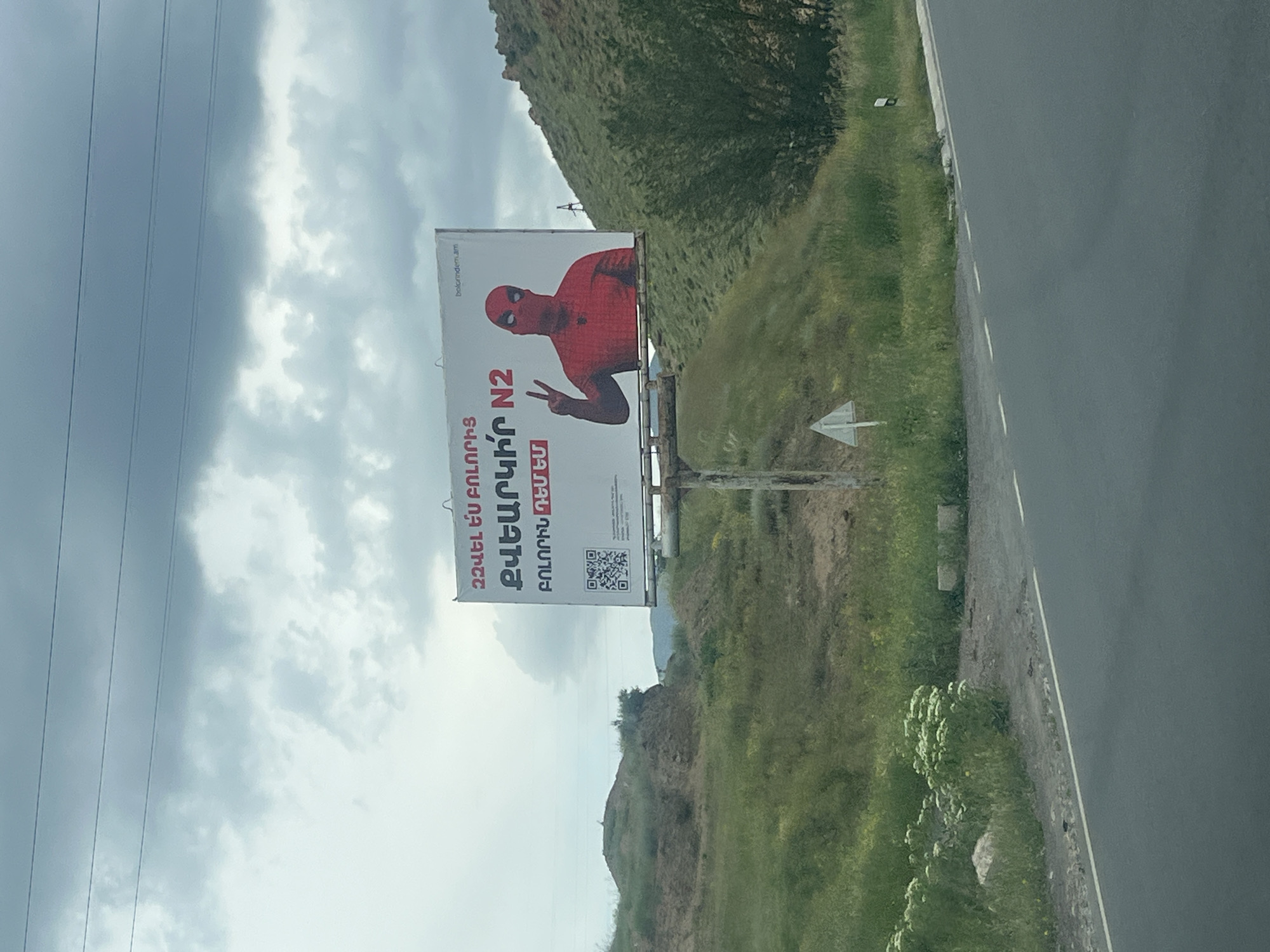 This full-shot image, captured in Chiva, Armenia, depicts a roadside billboard set against a backdrop of cloudy skies and green, hilly terrain. The billboard features a red and black masked character, strongly resembling Deadpool, making a peace sign with their left hand. Dominant red Armenian text on the billboard reads, "ԶԲԱՂՎԵԼ ԷՆ ԲՈԼՈՐԻՆ ԲԱՆԱԿԻՐ N2 ԲՈԼՈՐԻՆ ՈՒՇՔ," which roughly translates to "Engage everyone, Become a soldier N2, Attention to all." A smaller vertical website address, "bolorimdirm.am," is visible at the top right of the billboard, and a QR code is present at the bottom.
Multiple power lines stretch across the top left portion of the frame, leading towards the cloudy sky. To the right, a paved road with a white edge line runs alongside the grassy verge. A small white arrow sign points left on the ground near the road. The surrounding landscape includes green hills and some rocky patches. The lighting indicates it is daytime. The scene captures a moment along a rural or semi-rural road, featuring a prominent billboard with a pop-culture inspired advertisement, likely for a recruitment or awareness campaign given the text.
