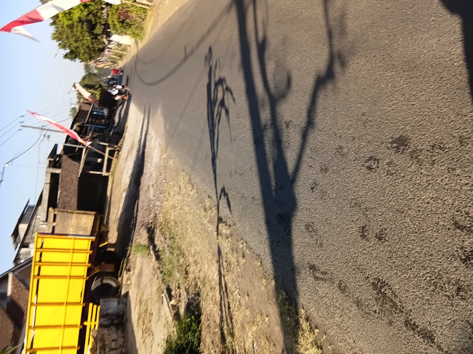 This image depicts a road scene in Tanggul Wetan, Indonesia. In the foreground, the asphalt road shows tire tracks and several dark, indistinct smudges that could be footprints or debris. The roadside to the left is dry and covered with sparse vegetation and dirt. A large, yellow truck with a metal cargo bed is parked on the left side of the road, partially obscuring some buildings. The buildings in the background appear to be residential or small commercial structures with tiled roofs. Indonesian flags, red and white, are visible strung on wires and poles along the road, indicating a local event or celebration. In the distance, a few people on motorcycles are traveling down the road, and more flags are visible. The sky is clear and blue, suggesting a sunny day. The overall impression is that of a typical rural or semi-urban street in Indonesia, possibly during a holiday.