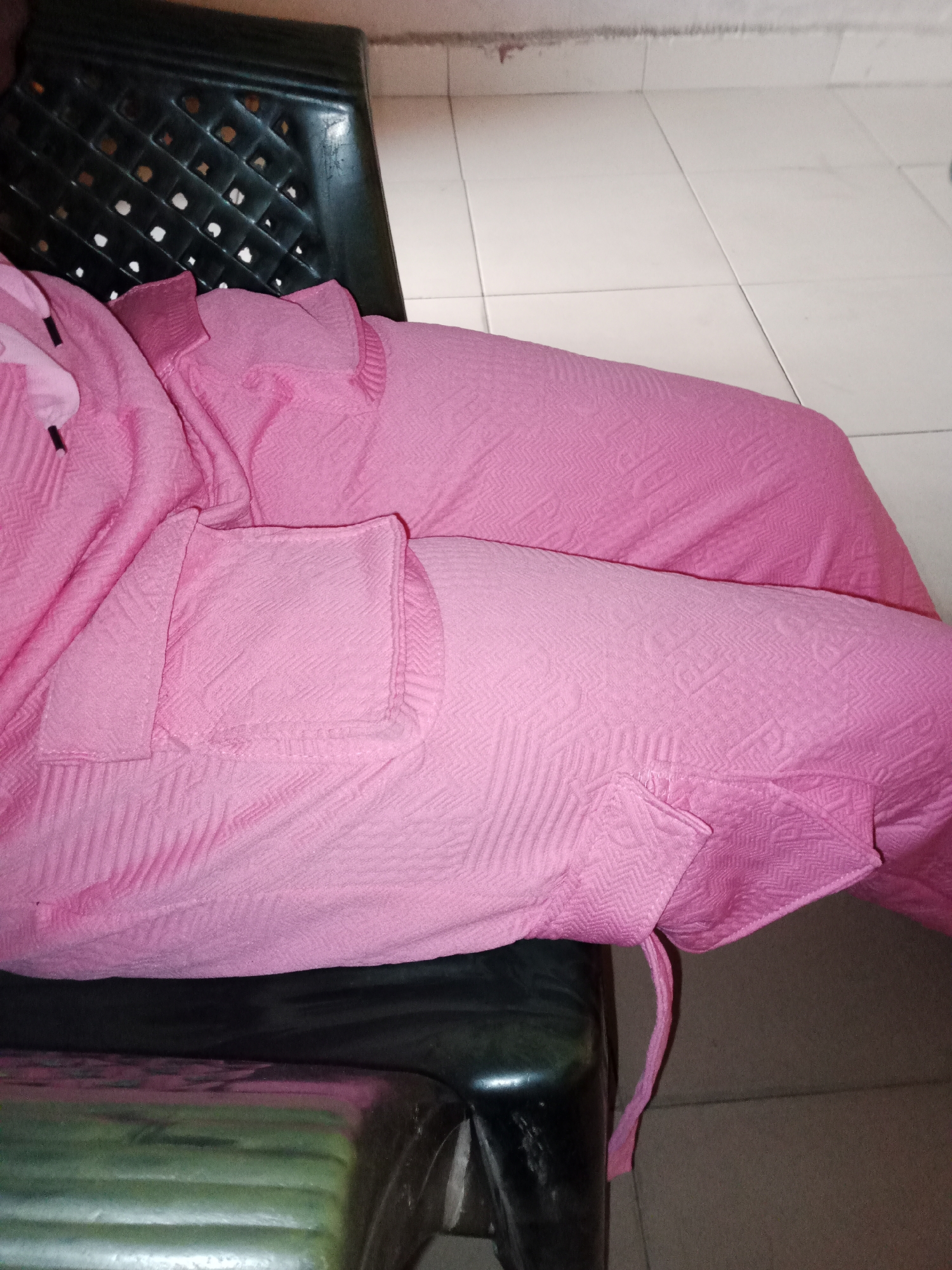 A person, likely female, is seated on a dark-colored chair with a woven back. They are wearing a pink, textured outfit, possibly a kaftan or abaya, which covers their legs and arms. The fabric has a subtle, repeating geometric pattern. The person's left arm is visible, bent at the elbow, and their hand is resting on their leg. The chair is positioned on a tiled floor, with a white and light brown tile pattern. A dark, possibly black, object is visible beneath the chair, appearing to be another piece of furniture or an accessory. The overall scene suggests an indoor setting, likely a home or a lounge area, in Jalingo, Nigeria.