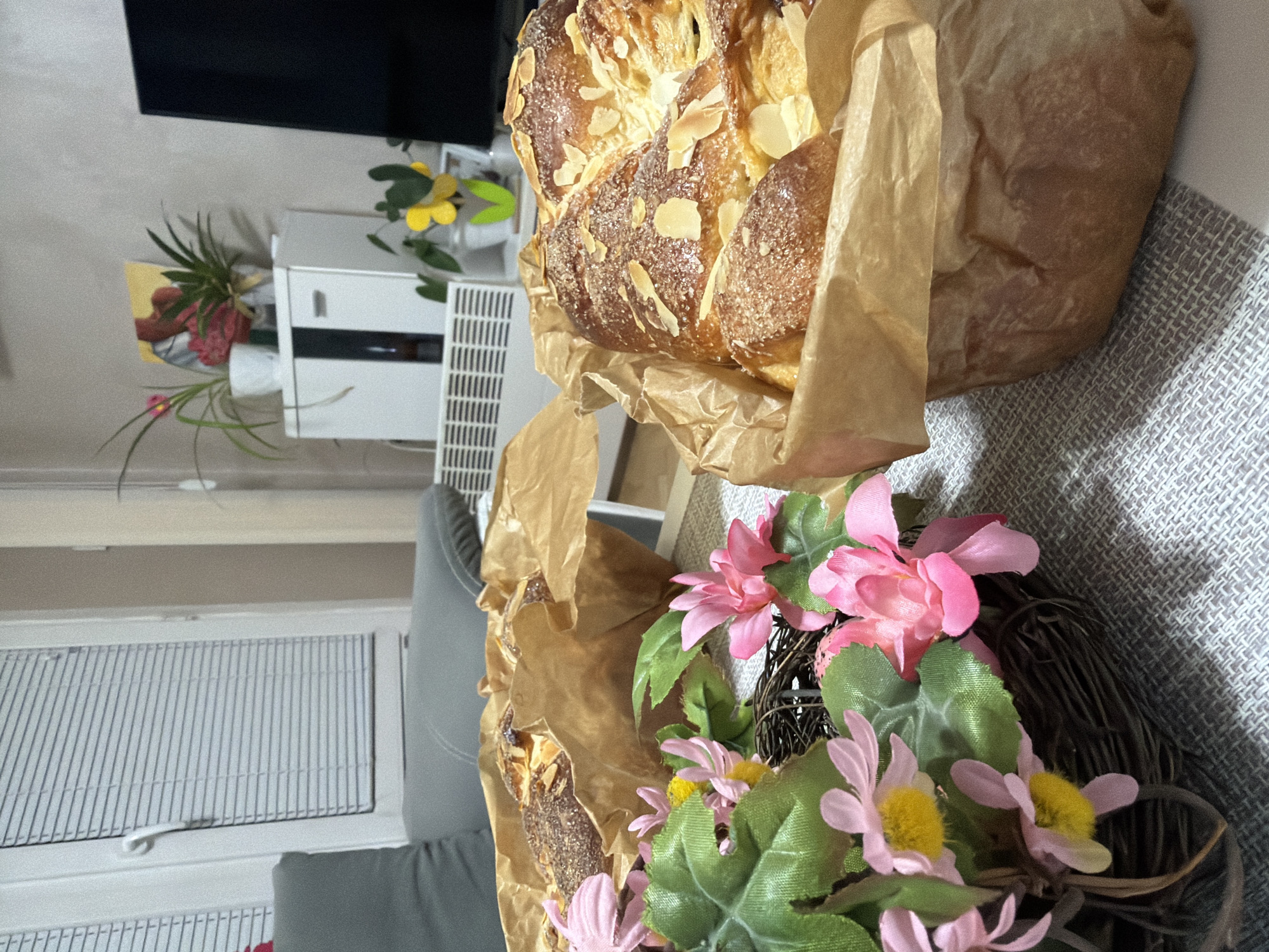 The image shows a loaf of bread topped with almonds in a brown paper bag, next to a floral wreath with pink and yellow flowers. The bread and wreath are resting on a gray textured surface. In the background, a window with blinds, a gray couch, a white cabinet with a TV on top, and potted plants are visible. The location is not identifiable as Plovdiv, Bulgaria.