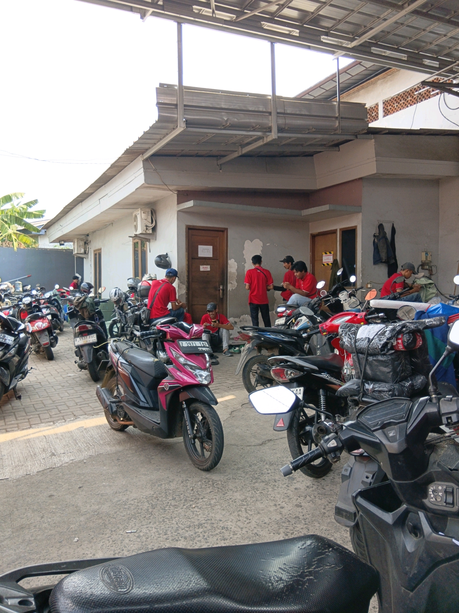 The image depicts a parking area in front of a building, likely a warehouse or distribution center, in Bekasi, Indonesia. The scene is dominated by a large number of parked motorcycles, suggesting a delivery or logistics operation. Several individuals, all wearing red t-shirts, are visible interacting within the parking lot. Some are seated on the ground or on motorcycles, while others stand and appear to be engaged in conversation or taking a break. The building itself is a simple, light-colored structure with a corrugated metal roof extension. The overall impression is one of an active, if informal, workplace.