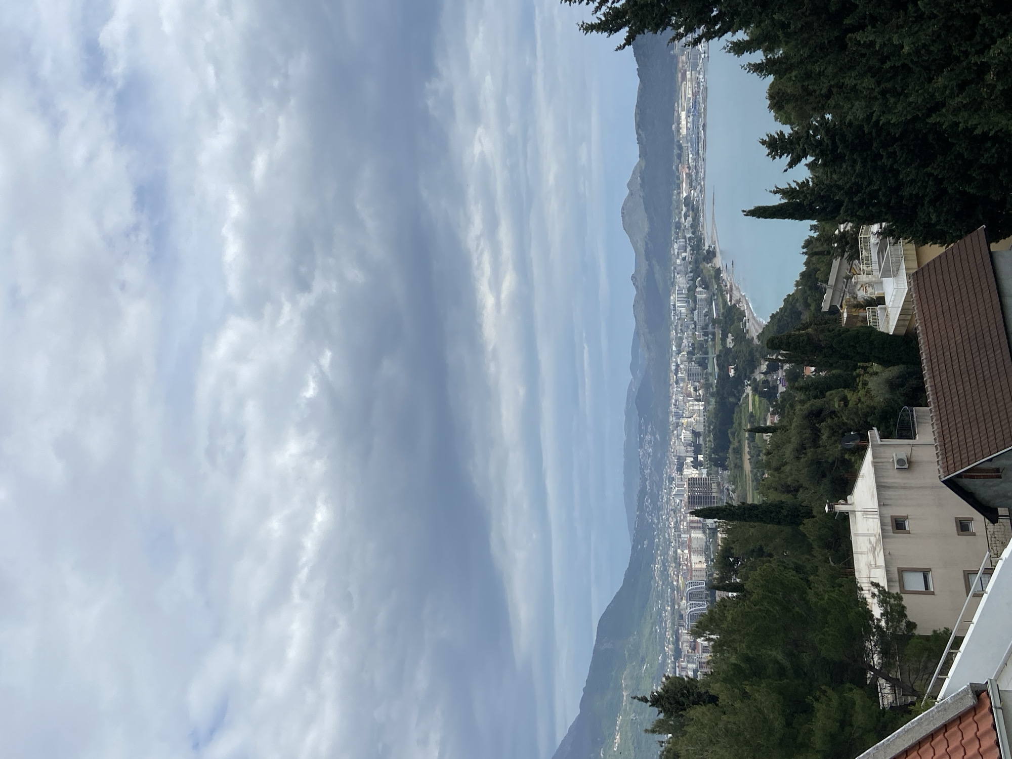 The image shows a panoramic view of Šušanj, Montenegro, from an elevated position. The sky is overcast with elongated cloud formations. In the middle ground, a coastal cityscape unfolds with buildings of varying heights interspersed with green areas of trees and grass. A narrow strip of coastline borders the city, next to a body of water. Mountains rise in the background, partially obscured by haze. In the foreground, the edge of a building with a brown roof and white walls is visible, along with evergreen trees along the top right.
