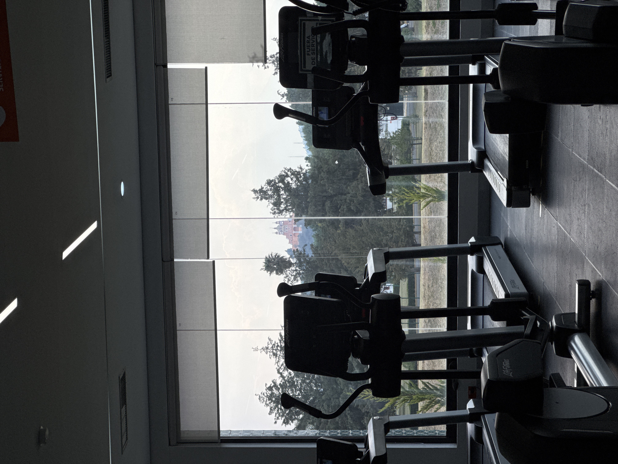 A gym is located in San Andrés Cholula, Mexico, with equipment facing a window overlooking a green space and a distant building. There are multiple elliptical machines with brand name "Life Fitness" visible. A sign that says "FUERA DE SERVICIO" in Spanish is also on one of the machines. The view through the window includes trees, a building in the distance.
