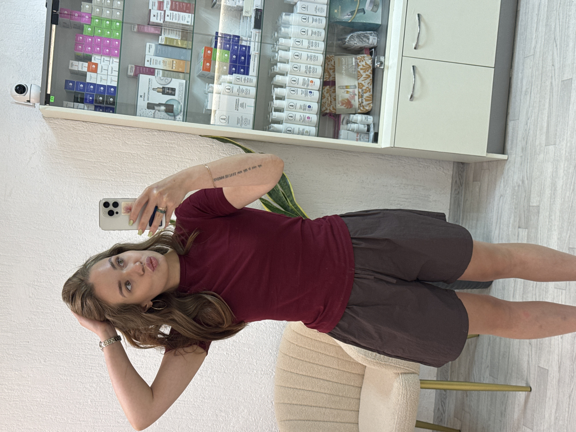 A young woman with long, wavy brown hair is taking a mirror selfie in what appears to be a salon or retail environment. She is wearing a maroon t-shirt and dark brown shorts. Her left arm is raised, with her hand touching her hair, and she is holding her phone in her right hand. The background includes a chair, a tall display cabinet filled with various cosmetic products, and a white wall. The floor is light-colored wood. A security camera is visible at the top of the display cabinet. The woman has light-colored nail polish on her fingers. The overall lighting suggests an indoor setting during the daytime. There is no specific information in the image to indicate the location is in the city of Chaikovsky, Russia.