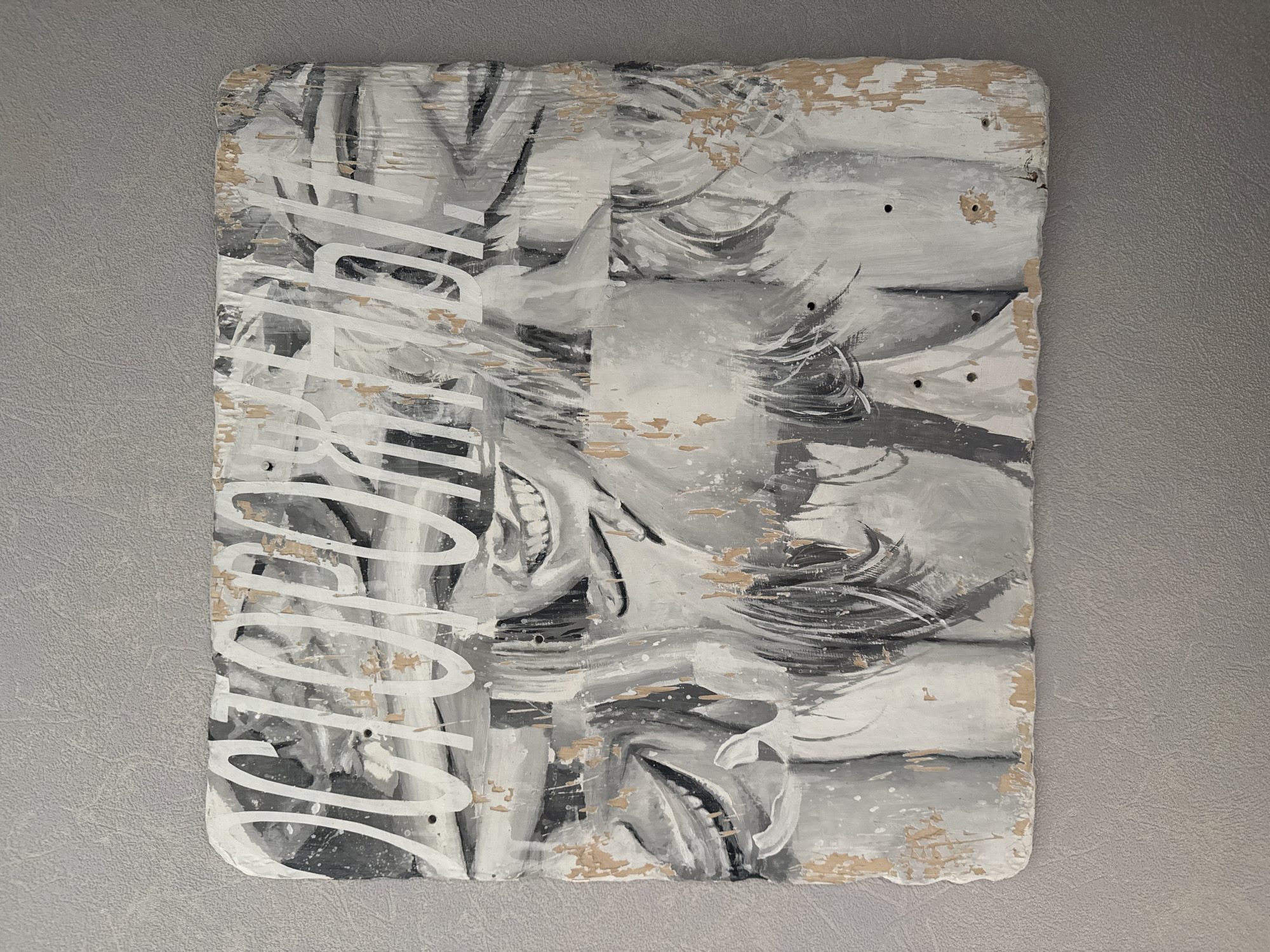An abstract mixed-media artwork, predominantly in grayscale, is presented on a distressed wooden panel. The panel, possibly a repurposed skateboard deck given the visible drill holes and uneven, chipped edges, serves as the canvas. The painting features intertwined forms rendered in black, white, and various shades of grey, with dynamic brushstrokes suggesting figures, possibly faces or bodies, and flowing elements like hair. Hints of open mouths with teeth are visible within the abstract composition.
A distinctive element is the stylized white text running vertically along the left side of the artwork. The letters are bold, rounded, and partially obscured by the painting style and overlapping forms. While challenging to read definitively, the shapes suggest letters like 'S', 'K', 'A', 'T', 'E', 'C', 'L', 'U', 'B', possibly spelling out "SKATE CLUB" or "SURF CLUB."
The artwork exhibits a rugged texture, with visible brushstrokes, paint splatters, and areas where the paint has chipped away, revealing the natural light brown/beige wood underneath. The entire piece is displayed against a textured, light grey wall. The visual context is an art piece, with no specific activity taking place other than its presentation. No particular time of day, weather, or overt emotions are discernible from the image. The location context provided is Moscow, Russia.