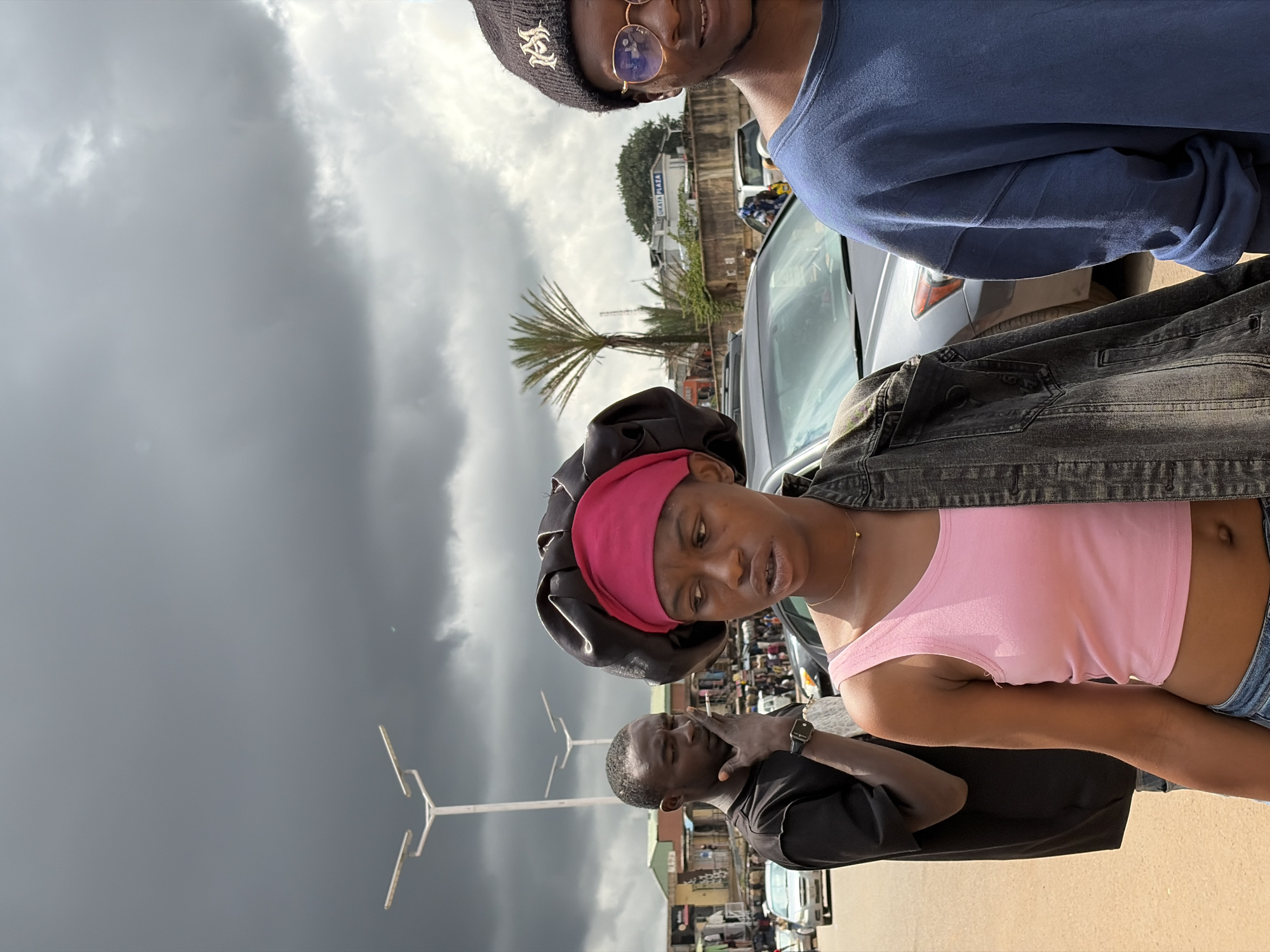 The image captures a street scene in Jos, Nigeria, under a cloudy sky. In the foreground, a young woman wearing a pink tank top, a black denim jacket draped over her shoulders, and a dark satin bonnet with a pink headscarf is looking to her right. She is wearing a delicate gold necklace. To her left, a young man in a black t-shirt and a watch on his left wrist is seen smoking a cigarette, with his hand resting near his mouth. Behind them, other individuals and vehicles are visible, suggesting a busy urban environment. Further in the background, buildings and streetlights line the street, and a palm tree stands tall against the overcast sky. The overall atmosphere is overcast, hinting at potential rain.