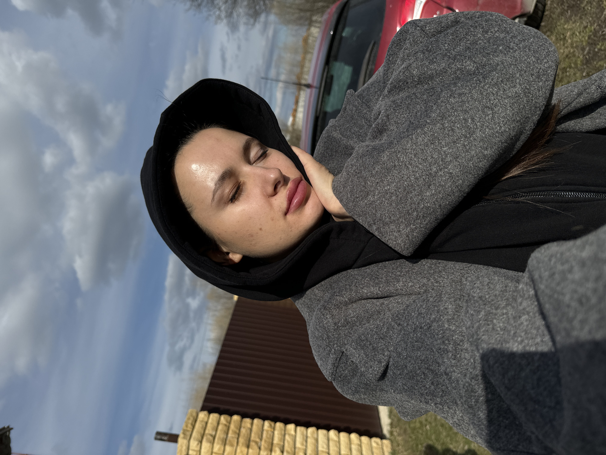 A woman with closed eyes and her hand on her face is wearing a black hoodie under a gray coat. A red vehicle is in the background. The environment includes a fence, grass, and a partly cloudy sky. The location appears to be outdoors, but the specific city is Unknown, Unknown.