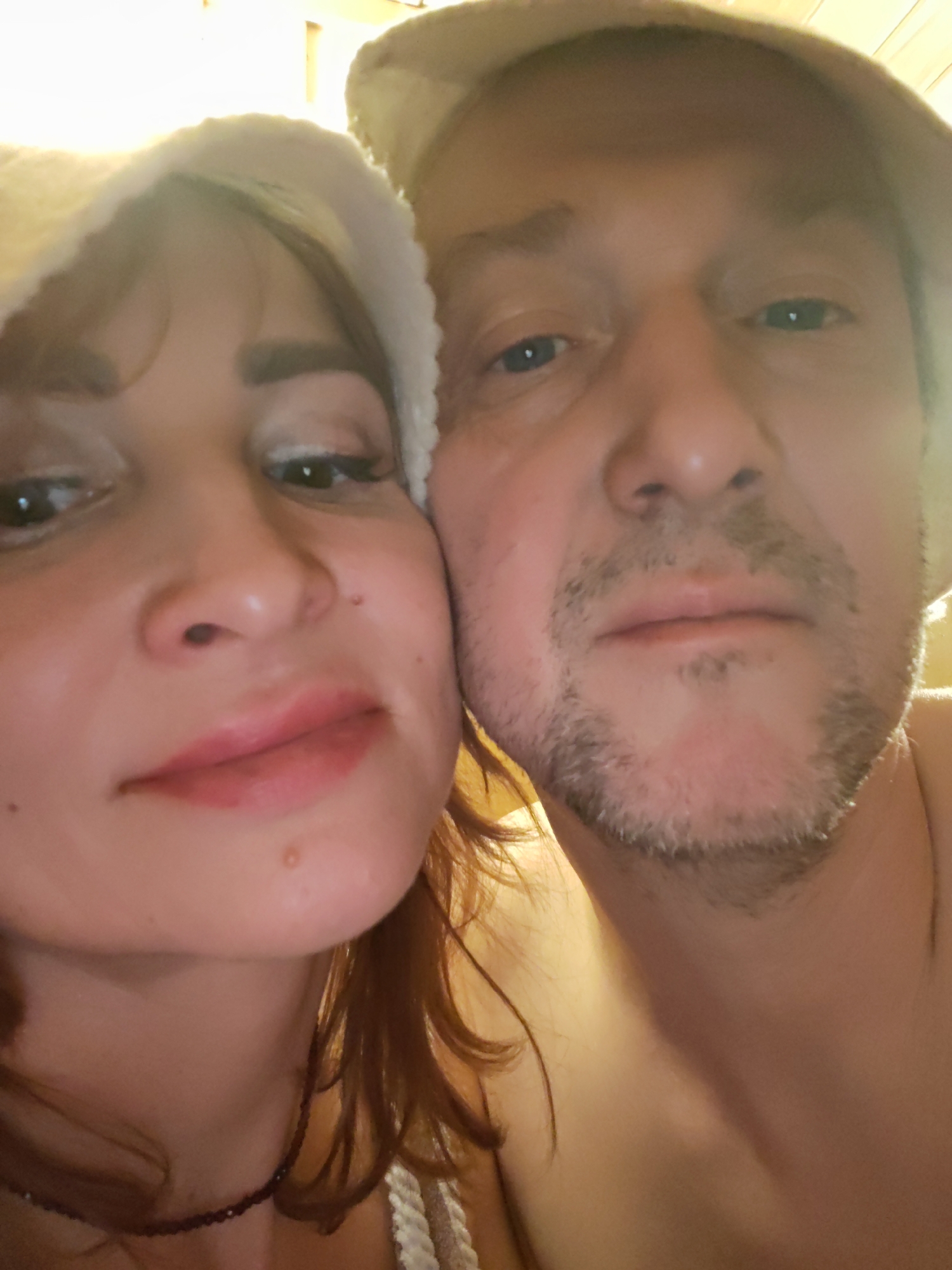 This image shows a close-up selfie of a man and a woman wearing what appear to be white sauna hats. The woman is on the left, with reddish-brown hair visible below her hat. She is wearing makeup, including eyeshadow and lipstick. The man is to the right of the woman and has stubble. They are close together, with their faces nearly touching. The background appears to be a warm, wooden-paneled surface, possibly indicating they are in a sauna or similar environment.