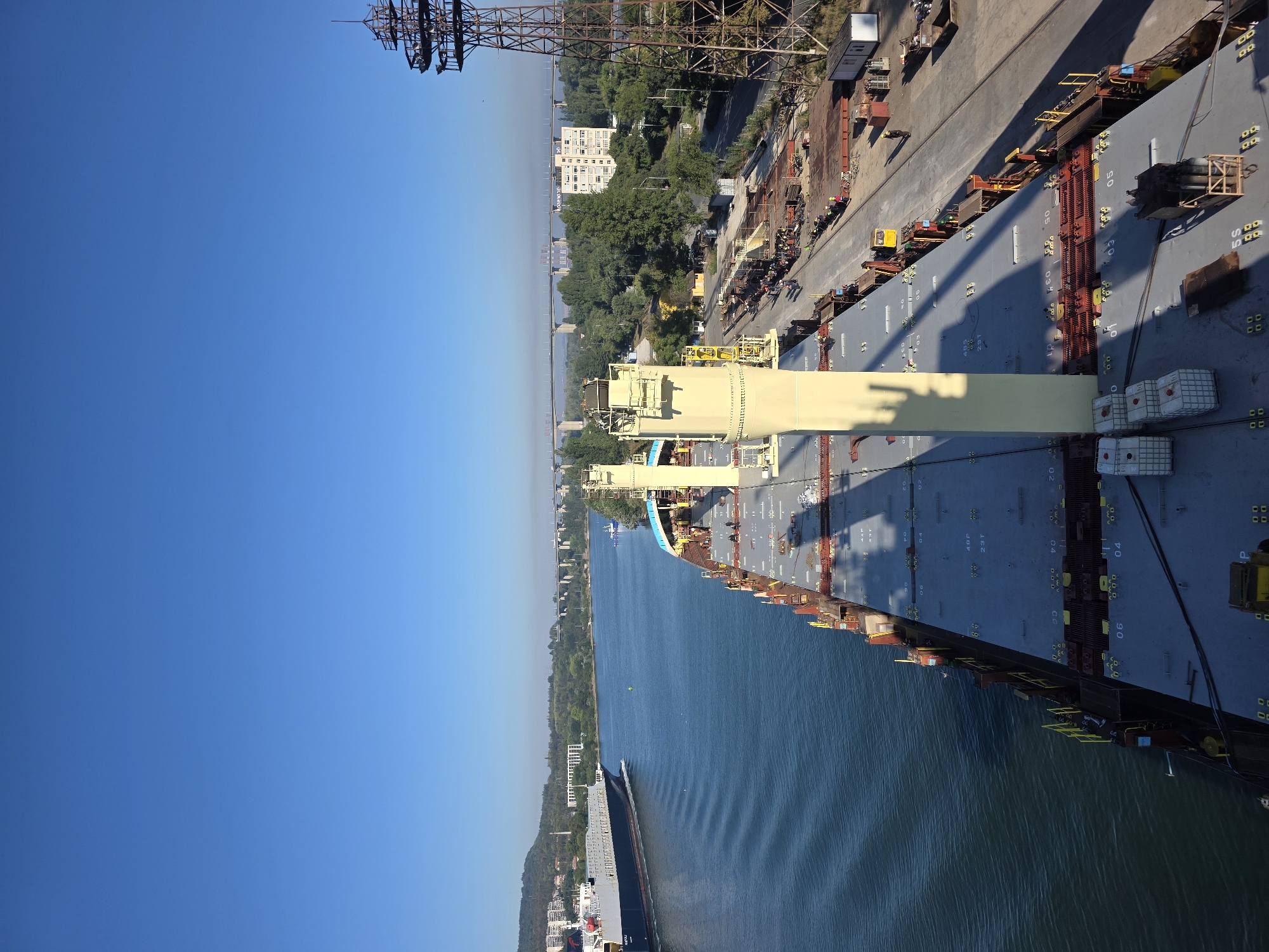 An aerial view shows a cargo ship docked at a port in Varna, Bulgaria, on a clear, sunny day. The ship's deck, covered with large, gray hatches marked with numbers and symbols, dominates the foreground. Tall, cream-colored structures and railings line the deck. To the left, a large blue and white cargo ship sails away from the viewer on a calm, blue sea, leaving a wake of ripples. The port area, visible to the right of the docked ship, features a large metal tower, railway tracks, piles of metal materials, and several small buildings. In the distance, a long bridge spans the water, connecting to a cityscape with buildings nestled amongst green trees and a hill. Several individuals, appearing as small figures, are visible on the dock, engaged in work. The overall scene suggests industrial activity within a coastal urban environment.