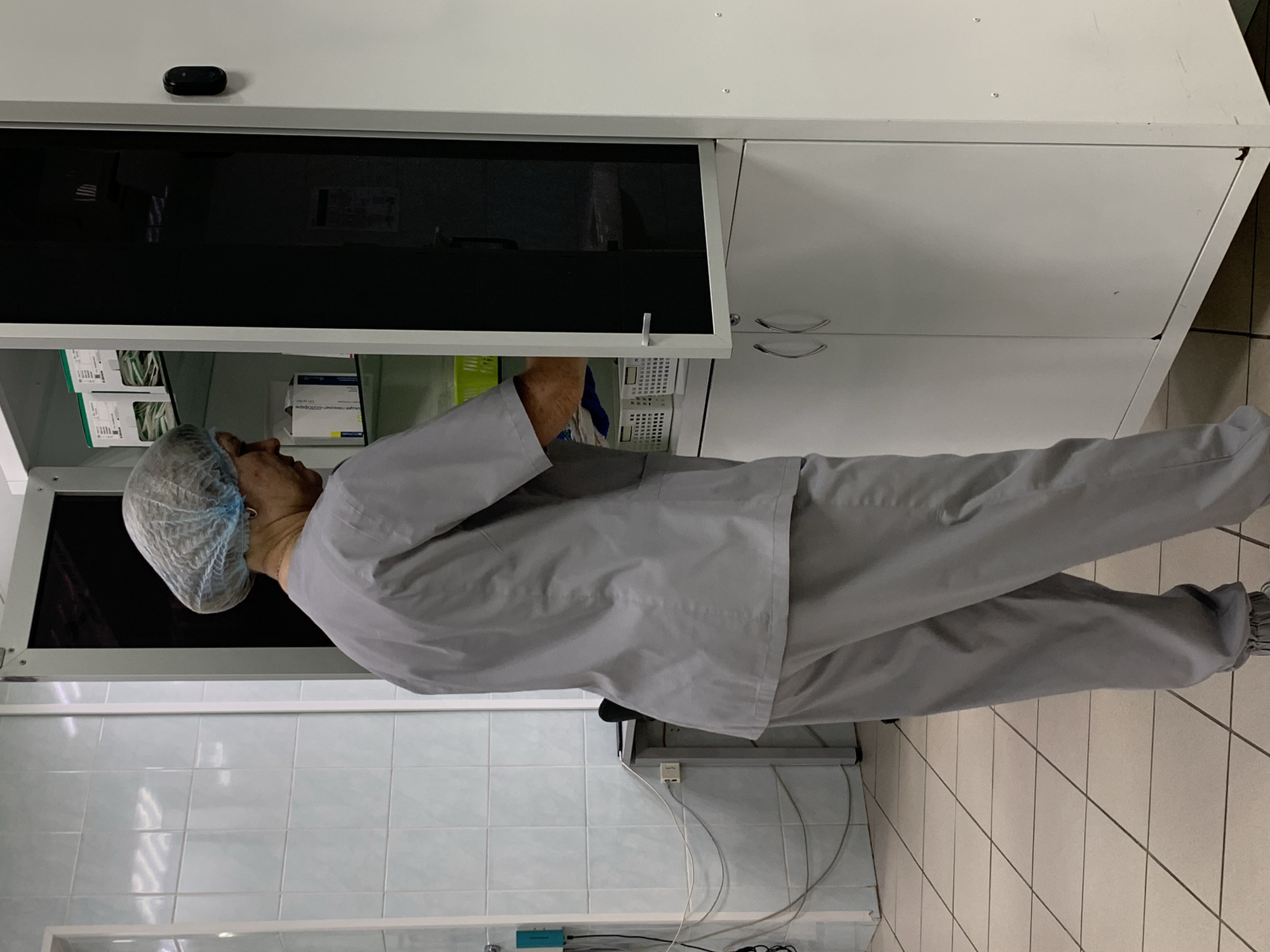 A person wearing a grey medical scrub suit and a blue hairnet is standing in a tiled room and reaching into an open cabinet with a dark glass door. The cabinet contains various medical supplies, including packaged items on shelves. The room appears to be a medical facility, possibly a hospital or clinic, located in the city of Chaikovsky, Russia. The floor is tiled with light-colored squares, and the walls are also tiled in a lighter shade. Electrical outlets and cables are visible on the wall behind the person. A white metal cabinet with two doors is positioned next to the cabinet being accessed.