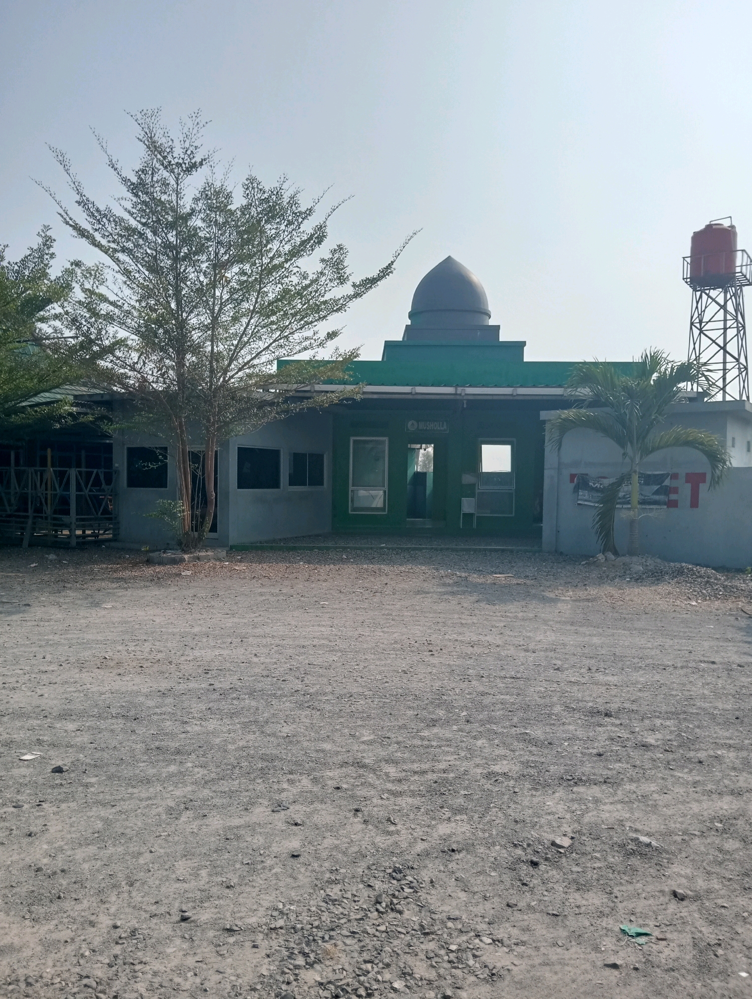 The image shows a view of a small building with a green facade and a dome, located in Wates, Indonesia. The building appears to be a musalla, as indicated by text on the facade. In front of the building, there is a dusty, gravel-covered area with some trees and vegetation. To the right of the building, there is a water tower. The sky is clear and bright. The scene is calm and serene, with no people visible in the frame.