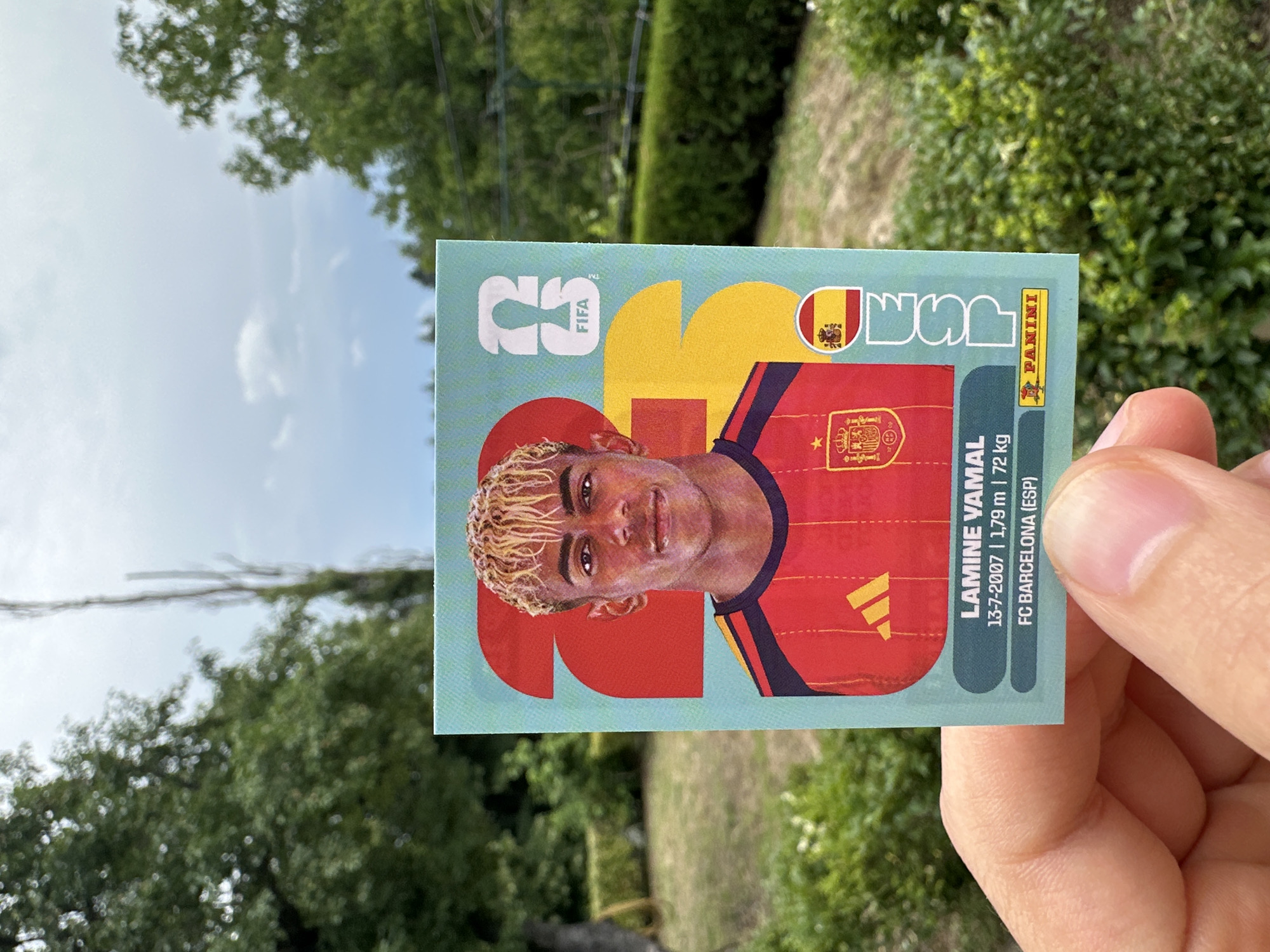 In Prisovo, Bulgaria, a person's fingers hold a Panini trading card featuring Lamine Yamal. The card displays the number "23" prominently, with Yamal, a young man with blond, spiky hair, wearing a red and yellow soccer jersey. Text on the card indicates his name, birthdate (13-7-2007), height (1.79m), weight (72kg), and club (FC Barcelona). The background shows lush green trees and a cloudy sky.