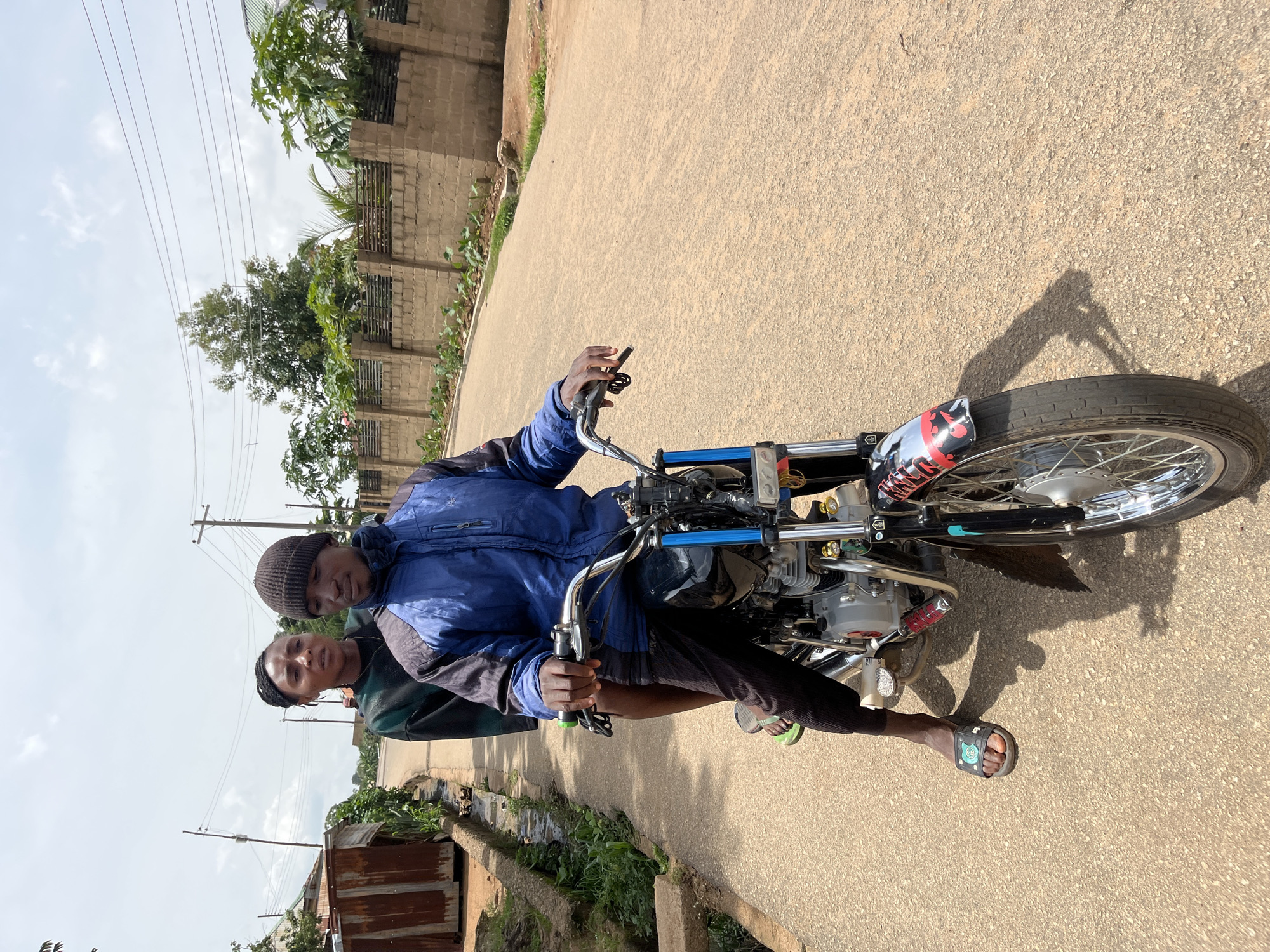 In Bukuru, Nigeria, two individuals ride a motorcycle on an asphalt road. The person in front, wearing a blue jacket and a dark beanie, operates the motorcycle. Behind them, a person wearing a green and black patterned top looks towards the camera with a concerned expression. The motorcycle is black with chrome accents and a red and silver decal on the front fender. The background shows a residential area with brick walls, vegetation, and utility poles under a cloudy sky. A small, rusty structure and a drainage ditch are visible to the left of the road.
