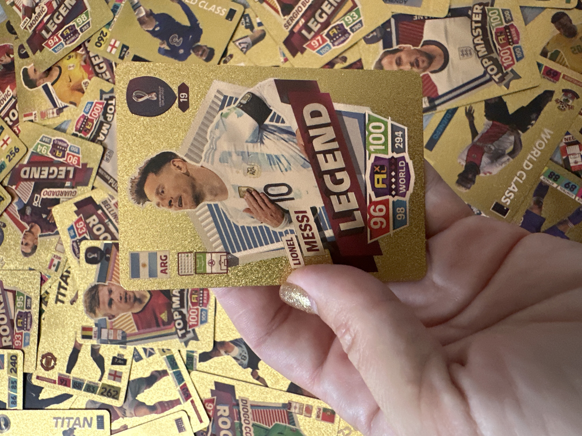 The image displays a close-up view of a hand holding a collectible soccer trading card featuring Lionel Messi. The card is gold-colored with "LEGEND" prominently displayed in large text. Messi is depicted in his Argentina national team kit, with the FIFA World Cup Qatar 2022 logo visible. Scattered around the main card are numerous other similar trading cards, many also gold-colored and featuring soccer players. Some of these cards show player names like "ROOKIE," "TITAN," "TOP MASTER," and "LEGEND." The background is cluttered with these cards, creating a layered effect. The image does not contain any information or visual cues to suggest a location in Samara, Russia.