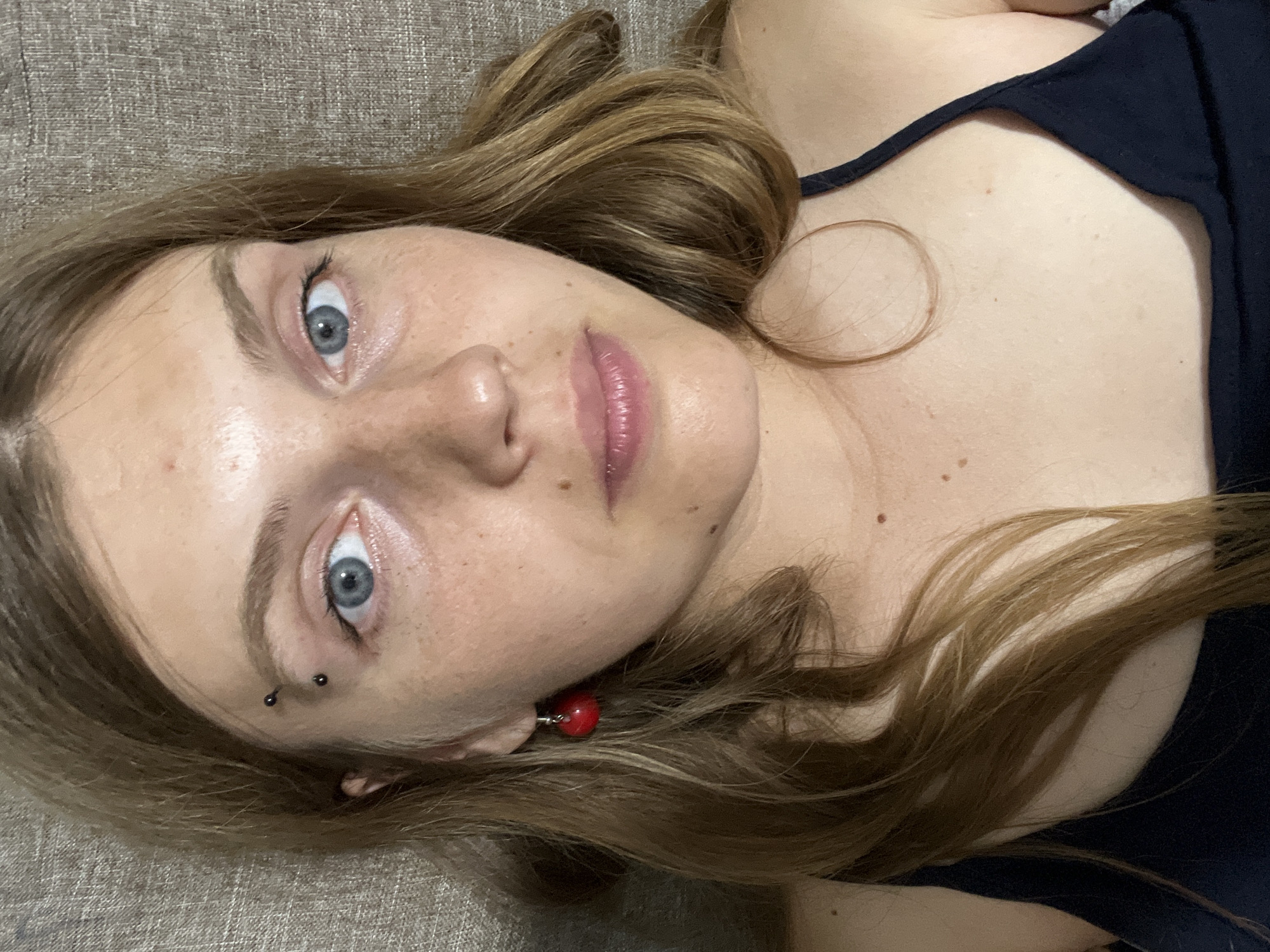 The image shows a close-up, overhead view of a young woman with light brown hair, blue eyes, and fair skin with freckles. She is wearing a dark blue tank top and a single red earring in her left ear. She also has a vertical eyebrow piercing on her left eyebrow. She is lying down on a textured, brownish-gray surface, possibly a couch or bed. The background is out of focus and appears to be a continuation of the same textured surface. There are no other people or objects visible in the image. The image does not provide any information about the location in the city of Сеніцкі сельскі Савет, Belarus.