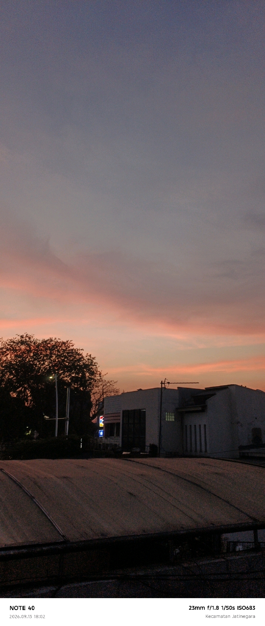 The image captures a dusky sky with hues of purple, grey, and vibrant orange and pink clouds. In the foreground, a dark, textured roof-like structure is visible, possibly industrial or a rooftop. A silhouette of a large, leafy tree stands to the left, illuminated by a streetlight. Behind the tree, a white building with a dark garage-like door and some windows is visible. The sky is filled with soft, wispy clouds, and the overall atmosphere is twilight. Text at the bottom of the image indicates "NOTE 40", the date "2026.09.13", and the time "18:02", along with camera settings "23mm f/1.8 1/50s ISO683" and the location "Kecamatan Jatinegara".