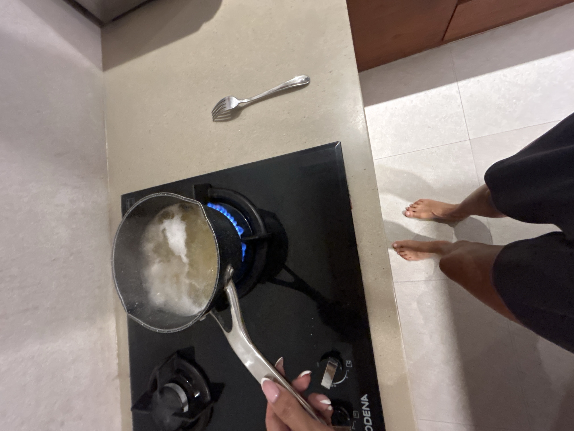 In a kitchen, a person's hand with manicured nails is holding a pot of boiling water on a gas stove. A fork lies to the right of the stove. The floor is tiled. The image is taken from a high angle, showing the person's feet wearing shorts. The background wall is textured and a countertop is visible. The scene appears to be located in Mengwi, Indonesia.