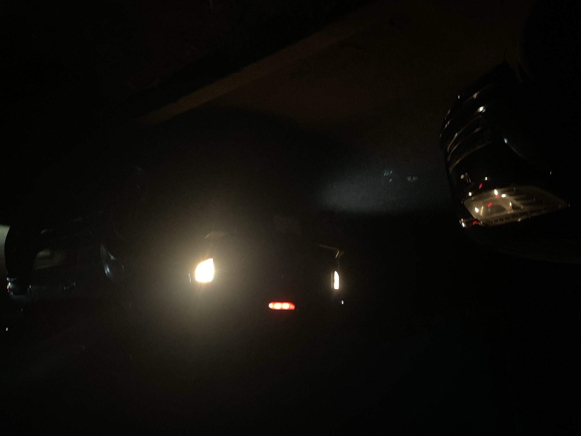 The image appears to be taken at night, possibly in an underpass or garage. A car with headlights and taillights is visible. Another vehicle with a partial view of the grille is on the right side of the frame. A possible ceiling or structure is visible in the upper background. No humans are directly visible. The location cannot be confirmed as Dutsen Alhaji, Nigeria.