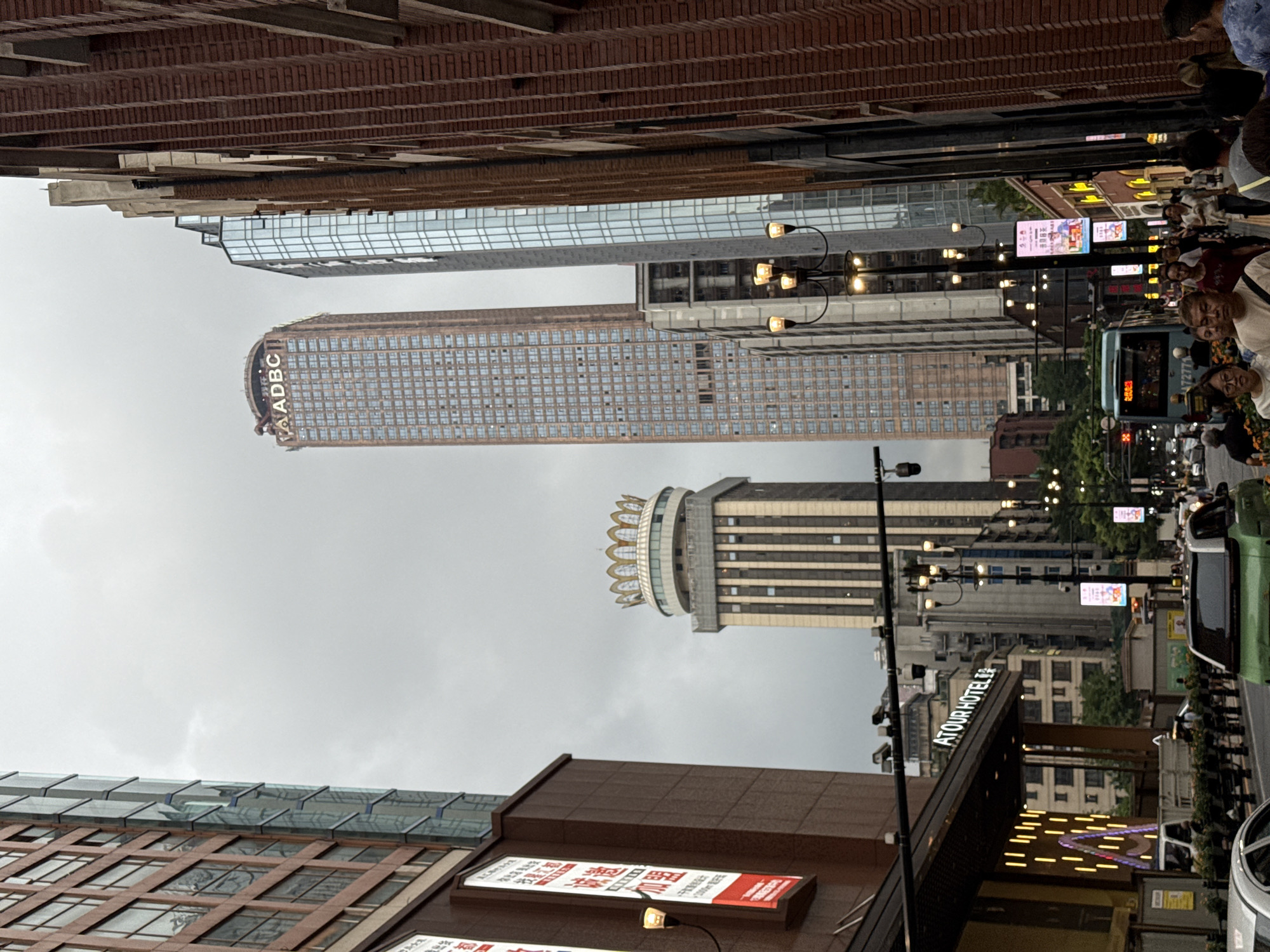 The image depicts a street scene in Yuzhong District, China, characterized by towering modern skyscrapers and a mix of pedestrians and vehicles. A tall, brown skyscraper with a grid of windows dominates the background, topped with the illuminated logo "ADBC". To its left, another building features a distinct crown-like structure. The foreground shows a bustling street with people walking along the sidewalk and cars, including a green vehicle, on the road. A bus with illuminated signage is also visible. Several streetlights with multiple lamps are positioned along the sidewalk, illuminating the path. The buildings are a mix of brick and glass facades, creating a dense urban environment under a cloudy sky. Signs in Chinese are present on some of the buildings. The scene suggests an active commercial area in a metropolitan setting.