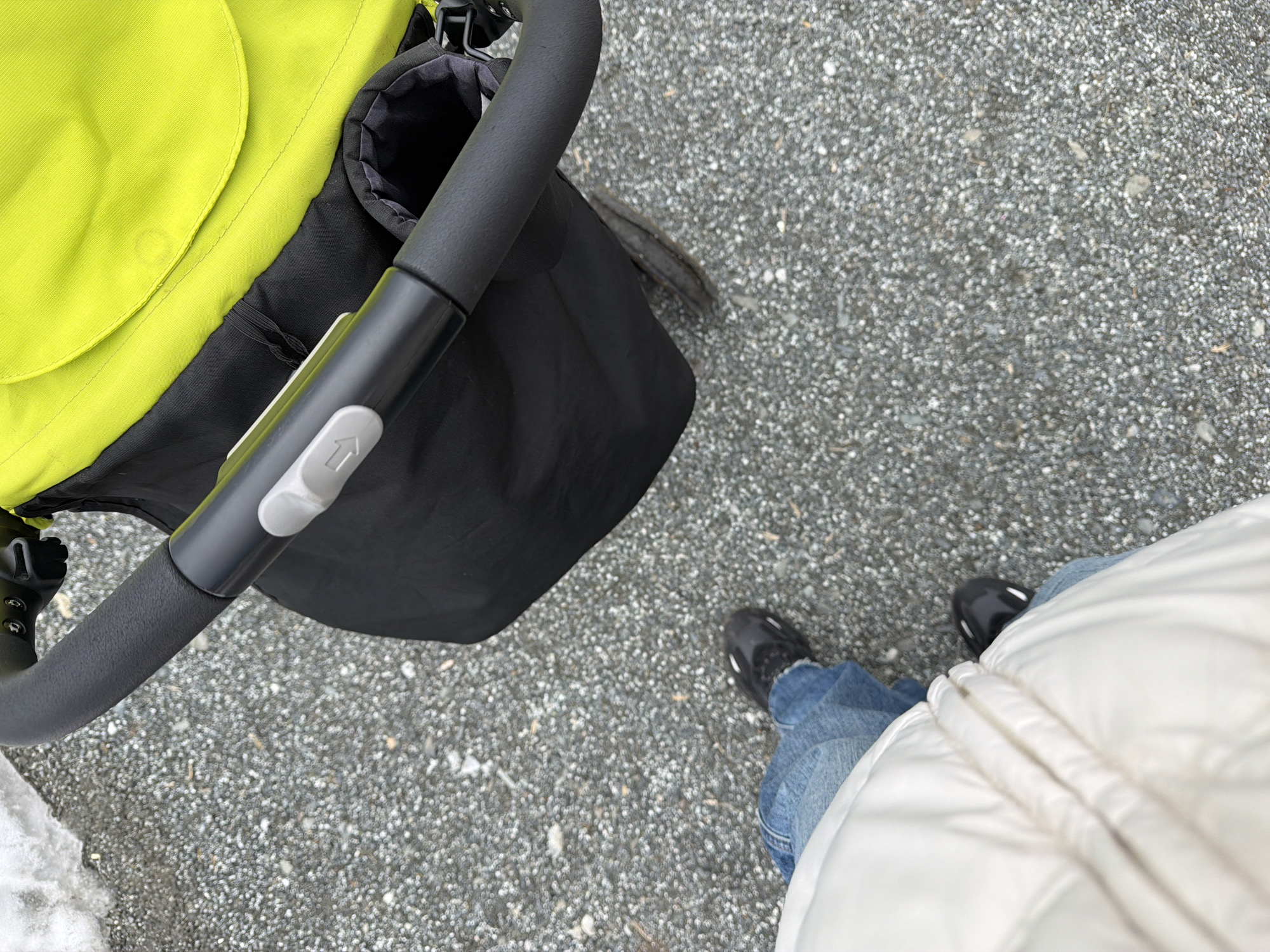 The image depicts an overhead view of a person standing on a gravel surface next to a baby stroller. The stroller features a neon green fabric section, a black storage bag, and a dark gray handlebar with a light gray button. The person is wearing blue jeans, black shoes, and a white puffy jacket. The background is a gravelly surface. The location is not discernible as being in Votkinsk, Russia; there are no recognizable landmarks.