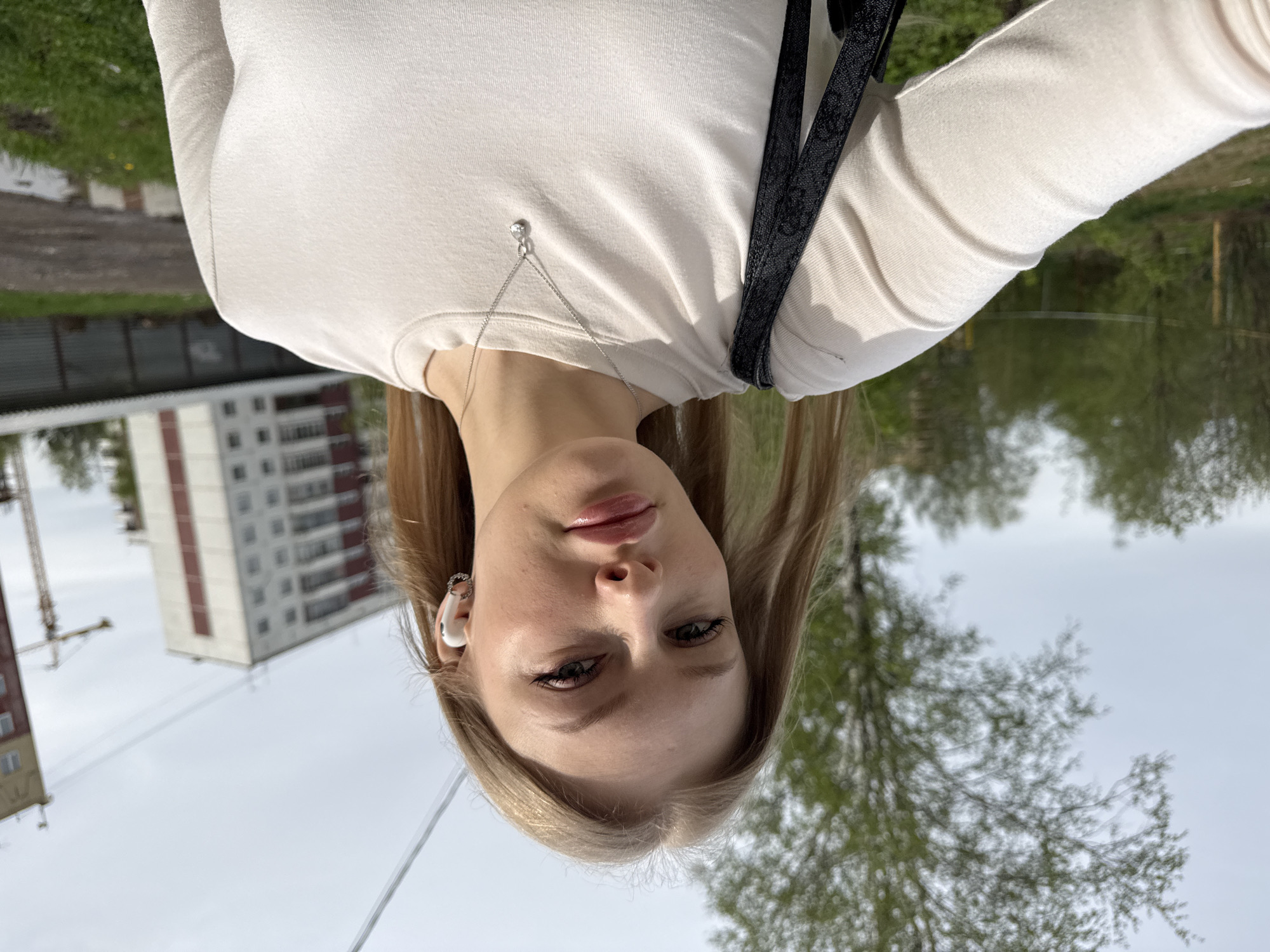 A young woman is taking a selfie outdoors in Perm, Russia. She has blonde hair, light skin, and is wearing a white long-sleeved shirt. She has a silver necklace with a small pendant, and a white wireless earbud is visible in her left ear. A black strap is visible over her shoulder. In the background, a tall apartment building with a red and white exterior is visible to the left. Green trees and a cloudy sky can be seen in the background.