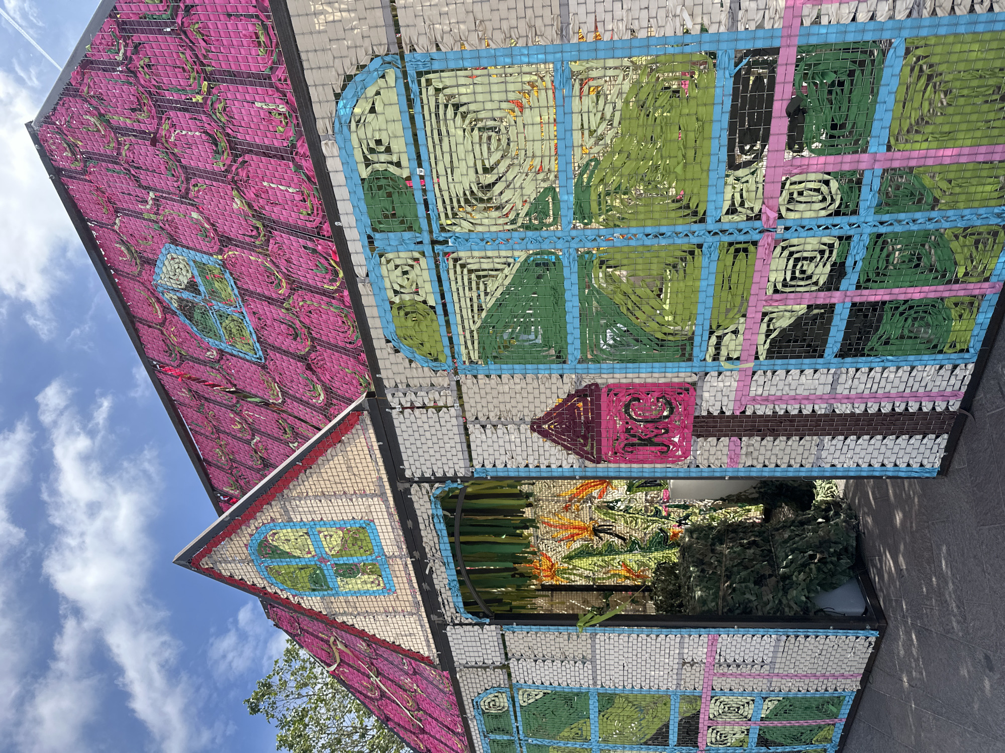 This image shows a colorful art installation in the form of a house, likely in London, United Kingdom, judging by the architectural style common in the city. The house is constructed with what appears to be woven material in a grid pattern, creating the structure. The "walls" are decorated with various colors, pinks, greens, and blues, and patterns representing windows and other architectural details. Inside the house, one can see plants and what appears to be a hidden space or a room within. The image is taken from a low angle, looking upwards toward the artwork, which suggests it is on display in a public space. The sky is visible in the background, indicating that it is outdoors. The house, with its whimsical design and bright colors, has an unusual, playful, and dreamlike atmosphere.