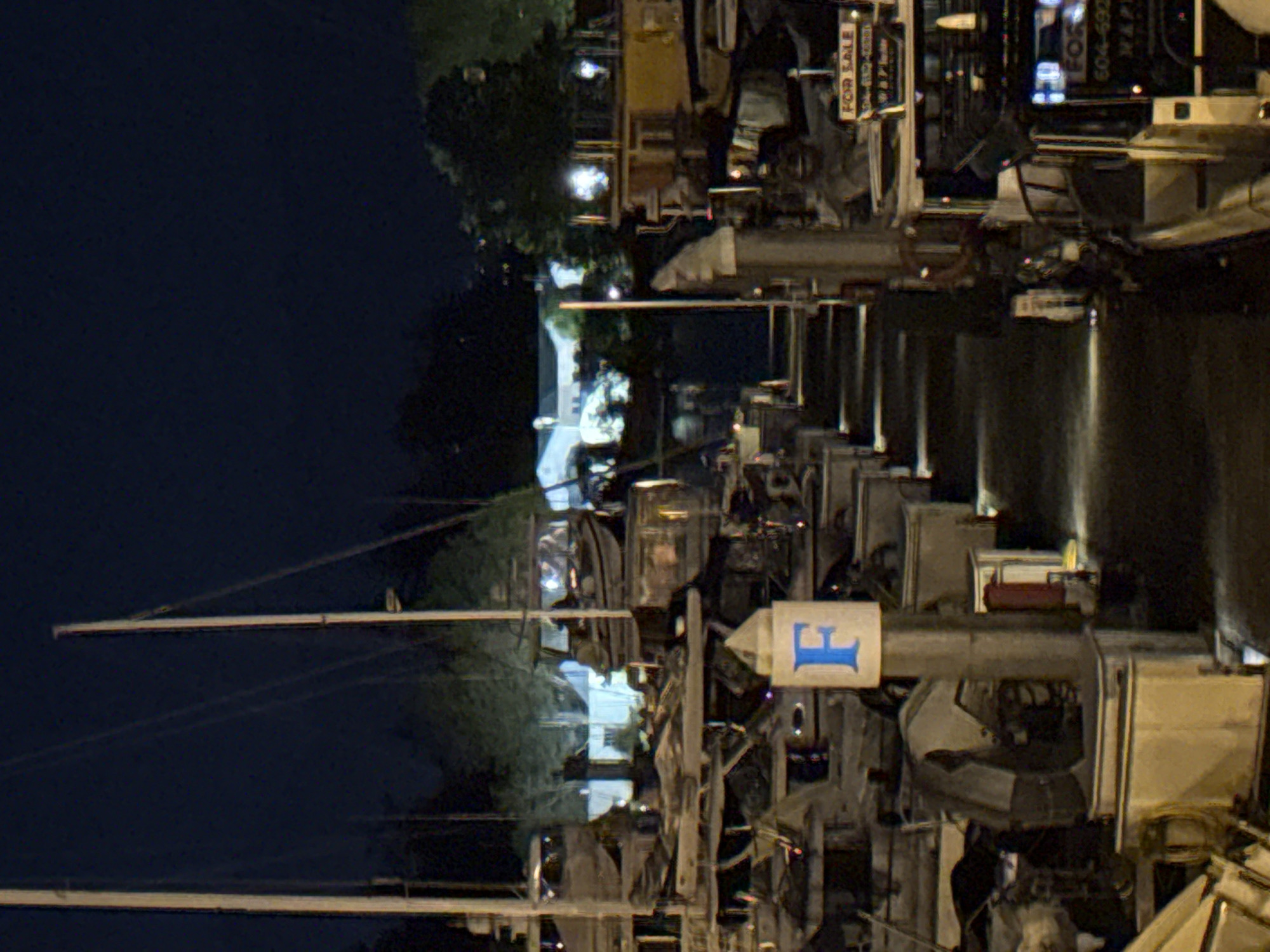 This night photograph shows a waterfront scene with docked sailboats and a marina in Vancouver, Canada. The masts of the sailboats extend into the dark sky. Docking infrastructure is visible, marked with lettered signs, indicating dock locations. Reflections of lights shimmer on the water's surface. Buildings and vegetation are visible along the edge of the water. Signage indicating the sale of items is also apparent.