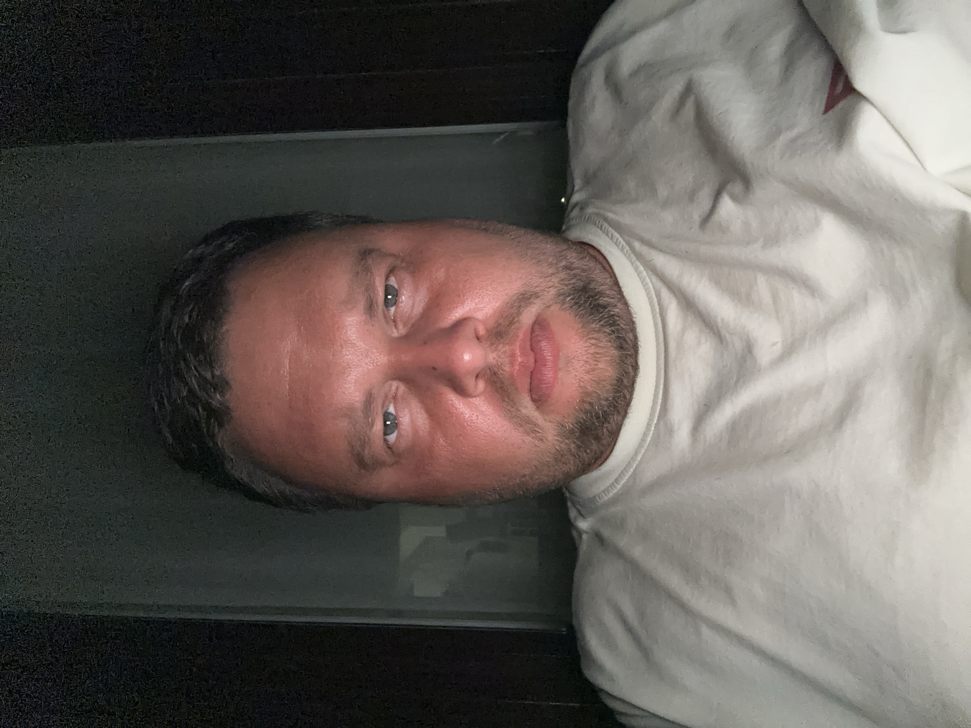A close-up, indoor portrait shot of a middle-aged man with fair skin, short dark hair, and a stubbly beard. He has light-colored eyes and is wearing a light-colored, crew neck shirt. The background consists of a dark surface, possibly a wall or a dark object, and a partially visible reflective or semi-transparent surface. The image does not contain enough information to identify the location as Sant Feliu de Guíxols, Spain or any specific actions or interactions.