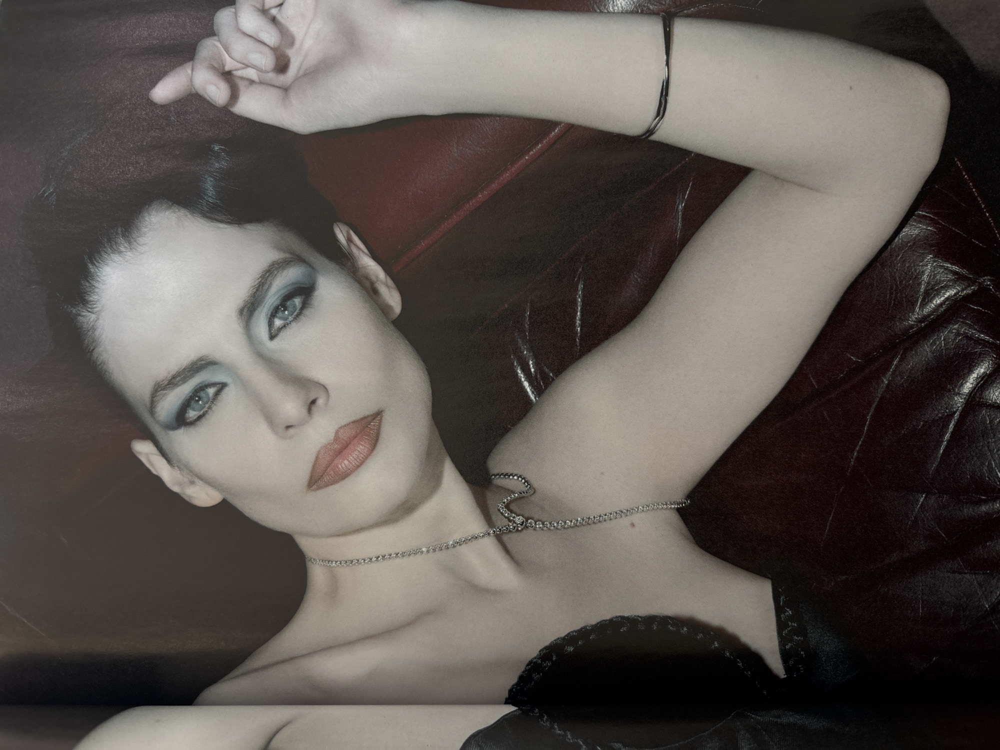 A pale-skinned woman with dark hair and blue eye makeup is lying on a dark brown leather surface. She is wearing a delicate silver necklace and a black top. Her left arm is bent and raised above her head, with a thin bracelet on her wrist. Her gaze is directed towards the viewer. The background appears to be a dark red leather surface.
