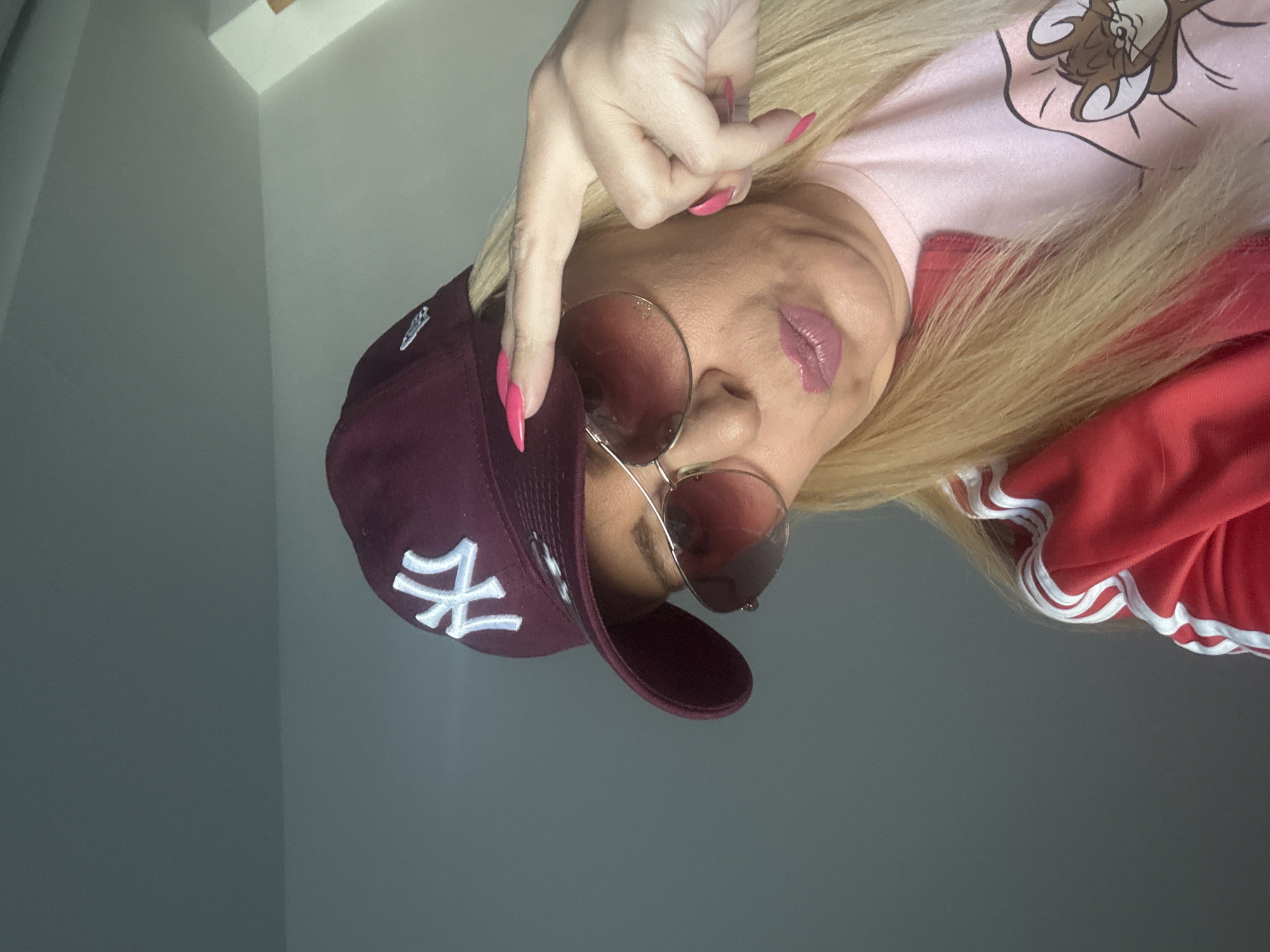 The image features a person's upper body and face, captured from a low-angle perspective looking slightly downwards into the camera. The individual has long, straight blonde hair that flows around their head. They are wearing a maroon baseball cap with the white 'NY' logo of the New York Yankees prominently displayed on the front. Large, aviator-style sunglasses with a reddish tint obscure their eyes, while their lips are adorned with light pink lipstick and are slightly parted.
A hand, with vibrant pink nail polish on the long, pointed nails, is raised into the frame, with the index finger pointing towards the viewer. The person is dressed in a light pink t-shirt featuring a cartoon character that resembles Taz from Looney Tunes on the chest. Over the t-shirt, they wear a red jacket or top with distinctive white stripes running along the sleeves. The background is a simple, light grey wall, with a visible corner and a white trim piece in the upper right, indicating an indoor setting. The overall impression is one of a casual and confident pose.