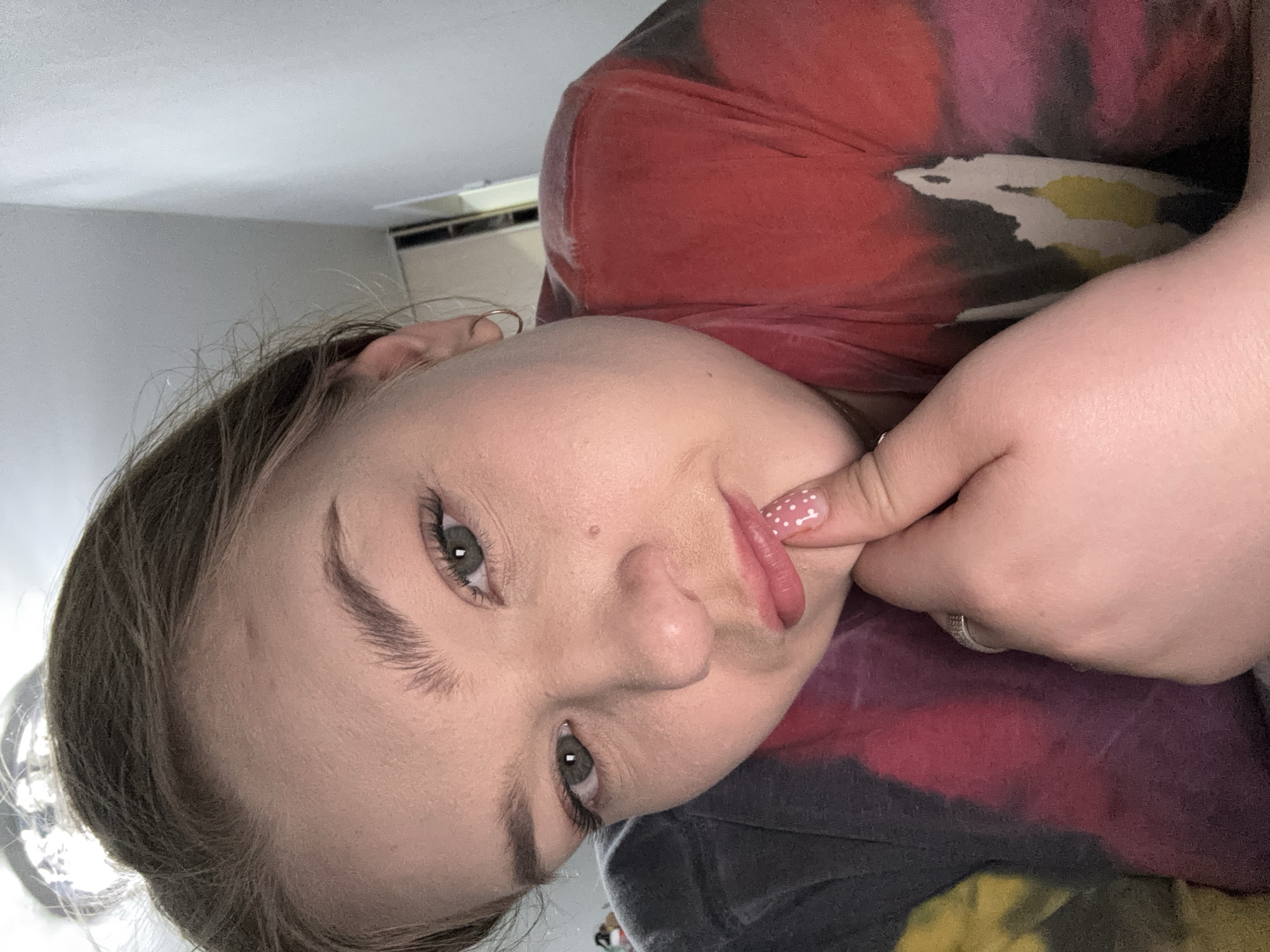 A person with light skin and gray eyes is posing for a selfie indoors. The person's face is tilted to the left and they are wearing a tie-dye shirt with red, pink, black, and yellow colors. The person is touching their lips with a finger that has white and pink polka dot nail polish. A ring is visible on the person's finger. An air conditioning unit is visible in the background, along with a white ceiling. The location in the city of Chaikovsky, Russia is not determinable from the image.