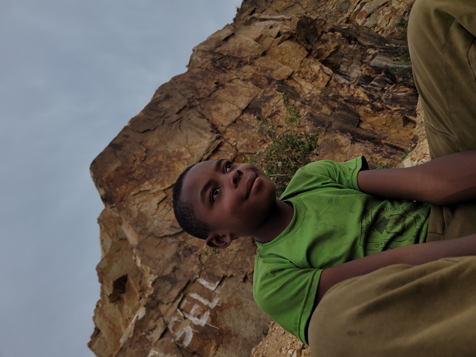 A medium, low-angle shot shows a young, dark-skinned boy sitting in front of a rocky cliff face. The boy is wearing a green t-shirt with a logo and tan pants. He is looking up and to the right with a slight smile. The cliff face behind him is made up of large, jagged rocks with some vegetation growing on it. The sky is overcast. The word "SELL" is written in white on the rocks at the bottom left of the image. The location is identified as Barakin Akawo, Nigeria.