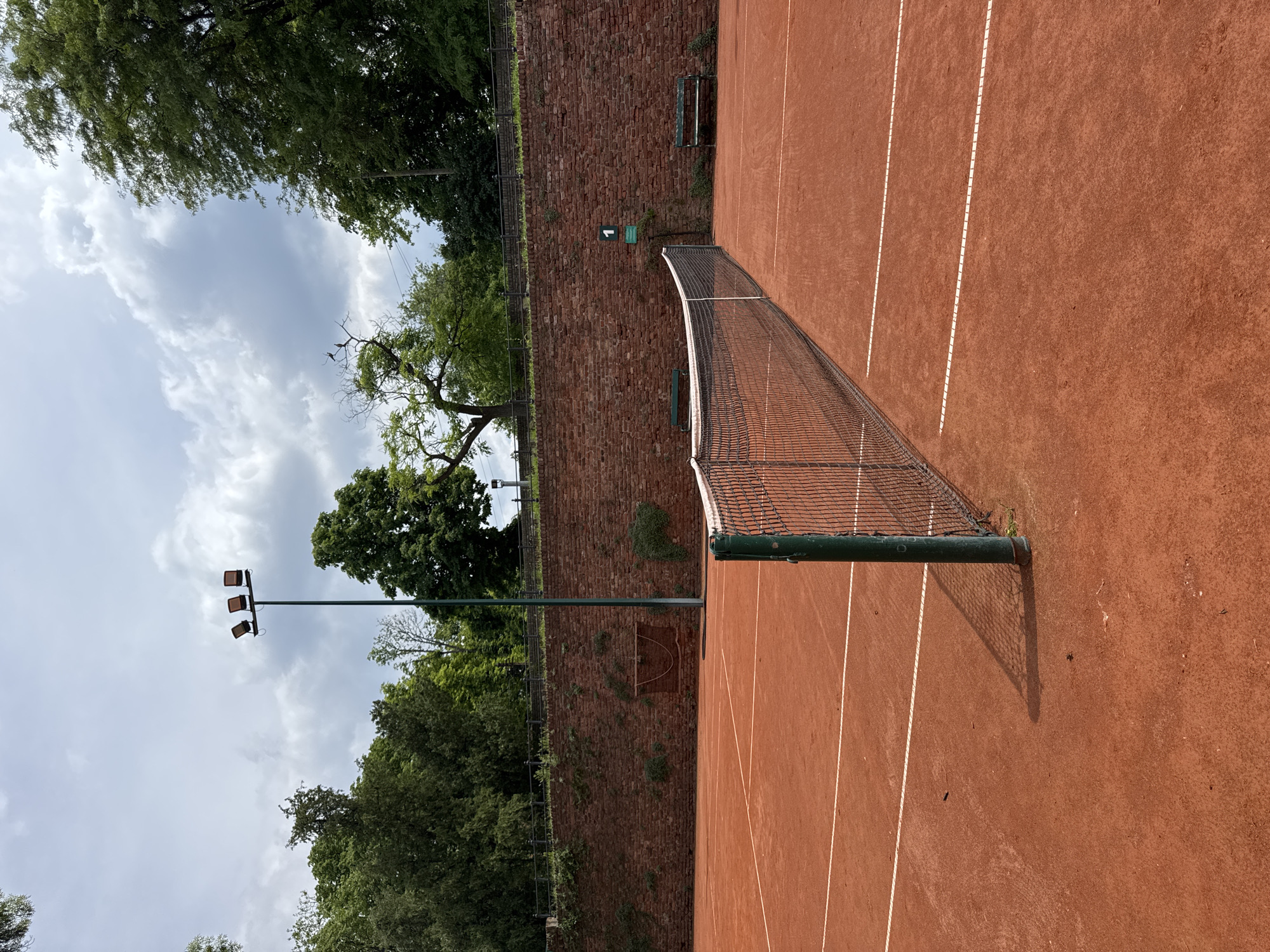 This is a photograph of a clay tennis court in Belgrade, Serbia. The court is surrounded by a brick wall on one side and trees overhead, creating a partially enclosed environment. A tennis net divides the court into two halves. The court's surface is a characteristic reddish-brown color, with white lines marking the boundaries. There is a lighting pole with lamps visible among the trees. A small bench is situated on the wall in the distance. The sky is overcast with scattered clouds.