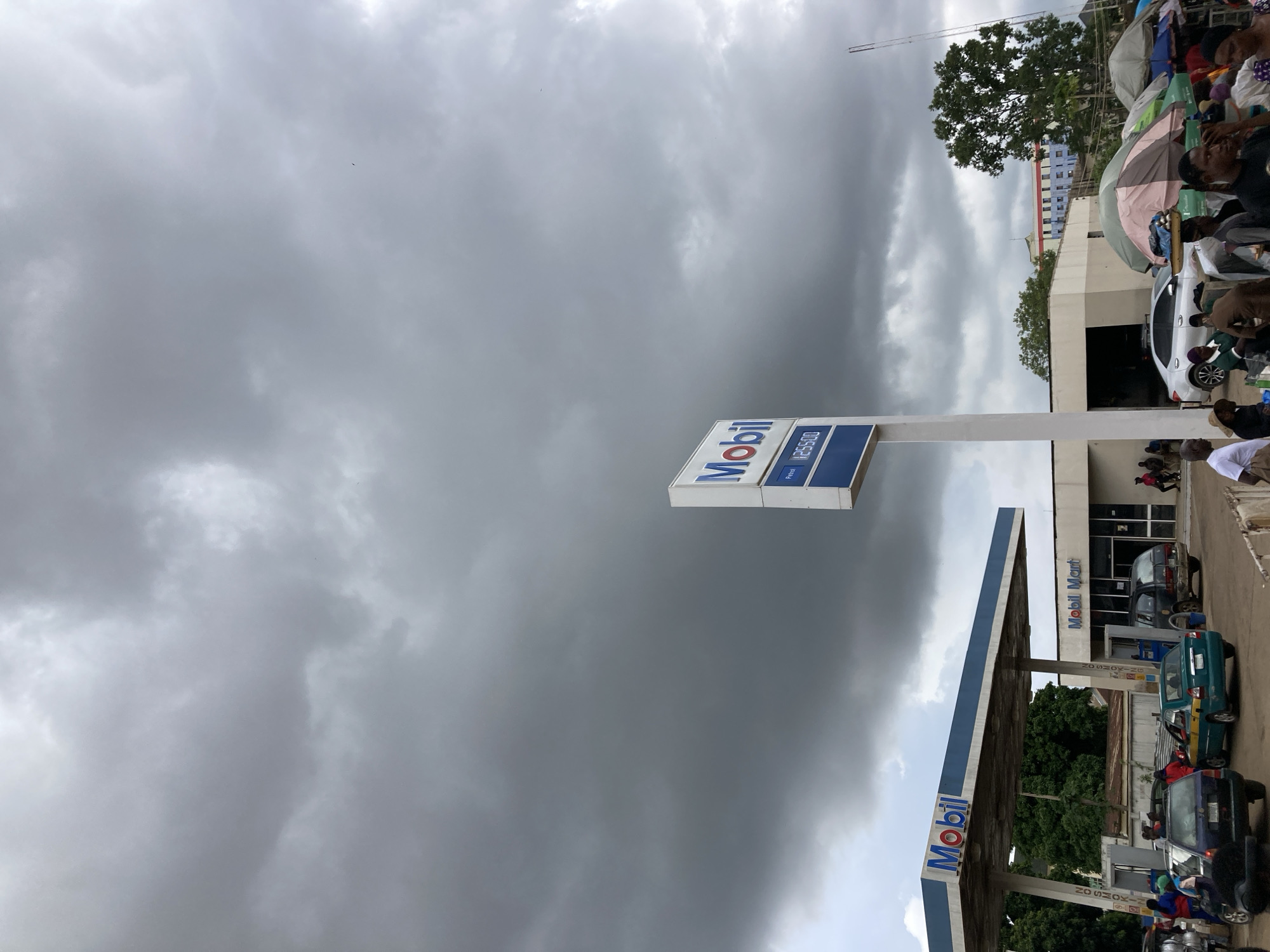 The image depicts a busy scene at a Mobil gas station in Jos, Nigeria. The sky is overcast with dark, dramatic clouds, suggesting an impending storm or heavy rain. The foreground features the prominent blue and white Mobil logo on a signpost and a gas pump canopy, with visible prices of P255.00 for petrol.
Underneath the canopy, several cars are lined up, indicating a period of activity and possibly a wait for fuel. People are visible around the station, some tending to vehicles, others gathered near stalls or shops adjacent to the gas station, creating a sense of a bustling marketplace. One of the buildings behind the gas station has "Mobil Mart" written on its facade.
The overall atmosphere appears somewhat chaotic due to the crowd and the weather, but there are no overt signs of distress or fear. The presence of people engaged in daily activities suggests a normal day, albeit under threatening skies. The location cues, such as the architecture and the "Mobil" branding, firmly place the scene in Nigeria. The time of day is unclear due to the heavy cloud cover, but it appears to be daytime.