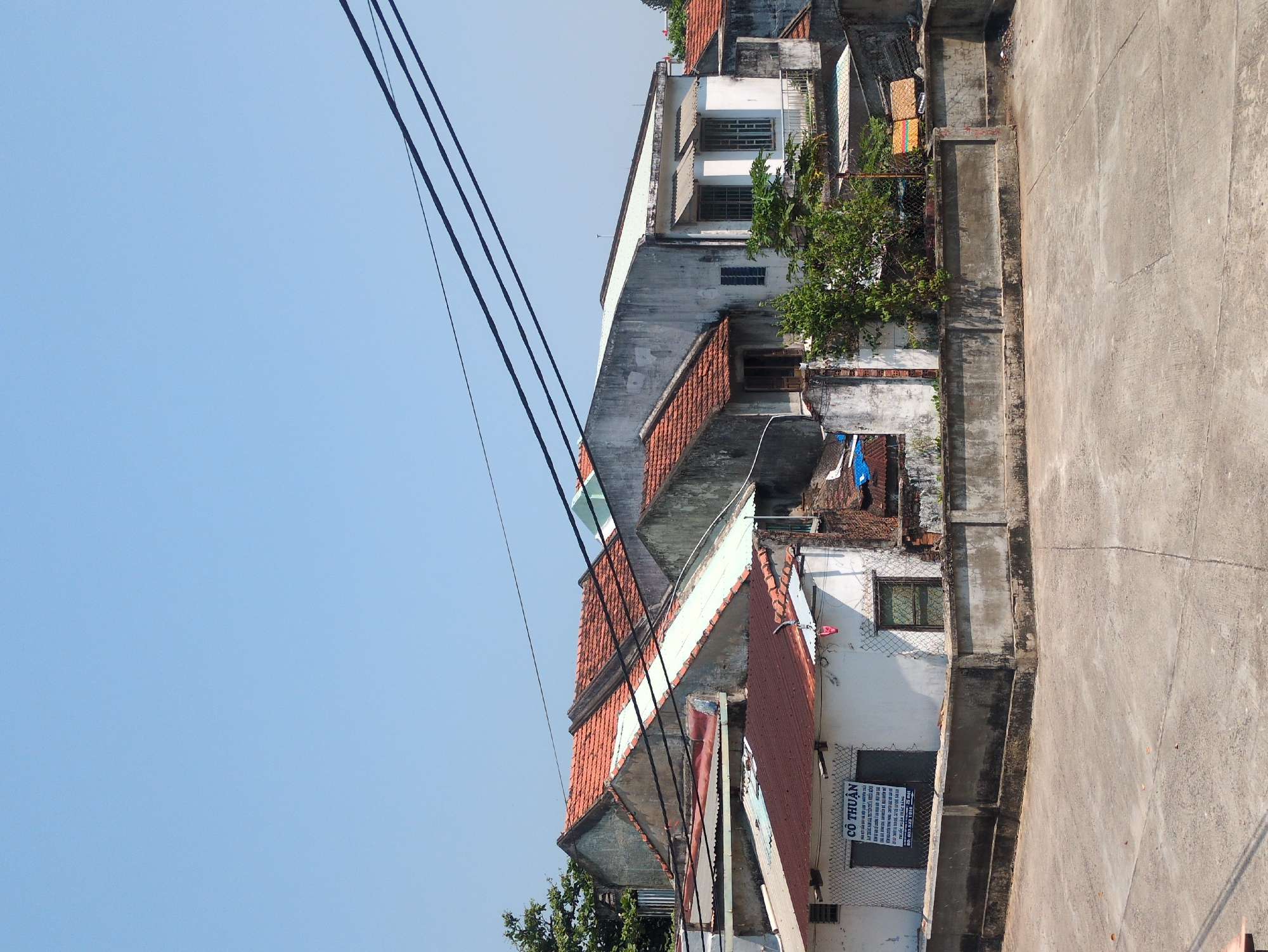 The image shows a street scene in Tuy Hòa, Vietnam. The buildings are old and built close together. The roofs are mostly orange tiles. Power lines run across the sky. The sidewalk is concrete. There are no people visible in the image. A sign is present.