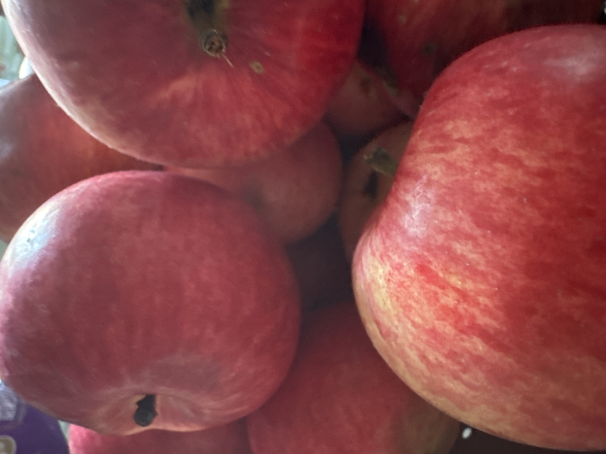 A close-up, overhead shot captures a collection of bright red apples piled together. The apples are ripe and appear to have a smooth, slightly waxy skin. Some have yellow or light red streaks, indicating variation in ripeness or variety. The lighting creates subtle shadows and highlights, emphasizing the roundness of the fruit. There are no visible people or actions, and the environment is not discernible beyond the immediate vicinity of the apples, which seem to be contained within a dark, possibly wooden, bowl or basket.