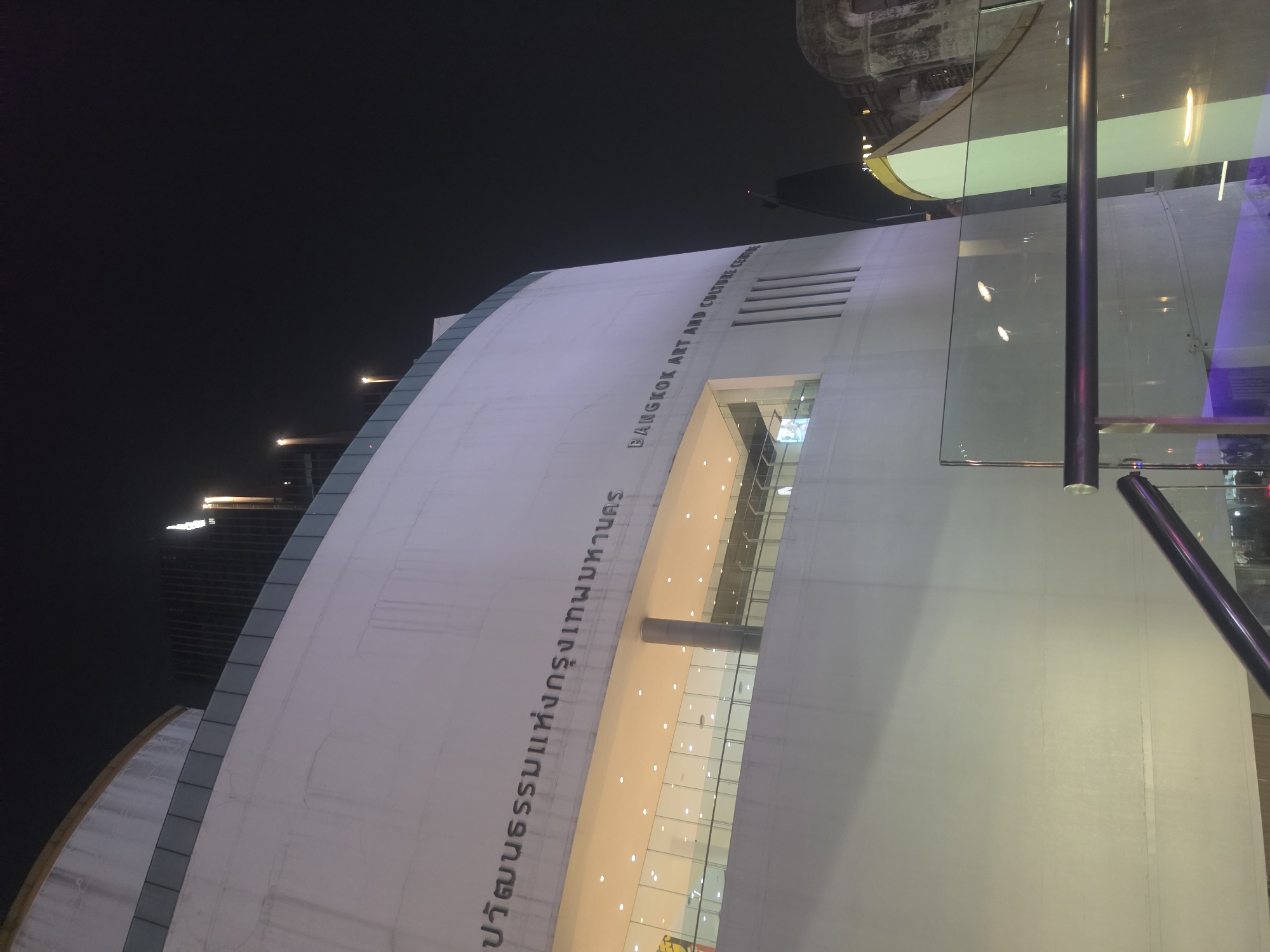 An exterior night shot captures the distinctive curved facade of a modern building, identified as the Bangkok Art and Culture Centre. The primary subject is the white, undulating structure, which features prominent horizontal text on its surface. To the left, the Thai inscription reads "หอศิลปวัฒนธรรมแห่งกรุงเทพมหานคร," while to the right, the English translation "BANGKOK ART AND CULTURE CENTRE" is visible.
The scene is set at night, with a dark, clear sky dominating the upper portion of the image. Artificial lights illuminate parts of the building's exterior and interior, the latter visible through large glass windows and entrances that reveal warmly lit spaces and structural elements. Reflections of these interior lights and surrounding structures are apparent in the glass panels, including a prominent dark vertical pole in the foreground. In the distant background, faint lights from other buildings are discernible against the dark sky. The overall architectural design suggests a contemporary cultural institution. No people or specific activities are visible, focusing solely on the building's imposing form and illuminated presence in the nighttime urban landscape of Bangkok.