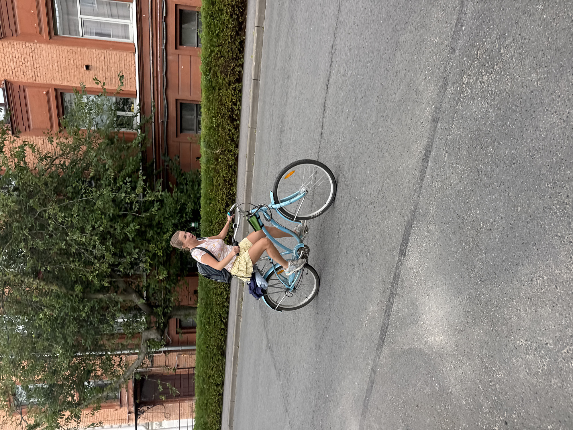 A woman rides a light blue bicycle on a paved road in the city of Ladozhskoe Ozero, Russia. She is wearing a light-colored t-shirt with a floral pattern and a yellow skirt. A dark backpack is on her back, and a blue jacket is tied to the rear rack of her bicycle. The bicycle has white handlebars and a light-colored seat. The woman is in motion, pedaling the bicycle. The background consists of a red brick building with windows and a thick green hedge. The road is asphalt and appears to be in good condition. The lighting suggests it is daytime.