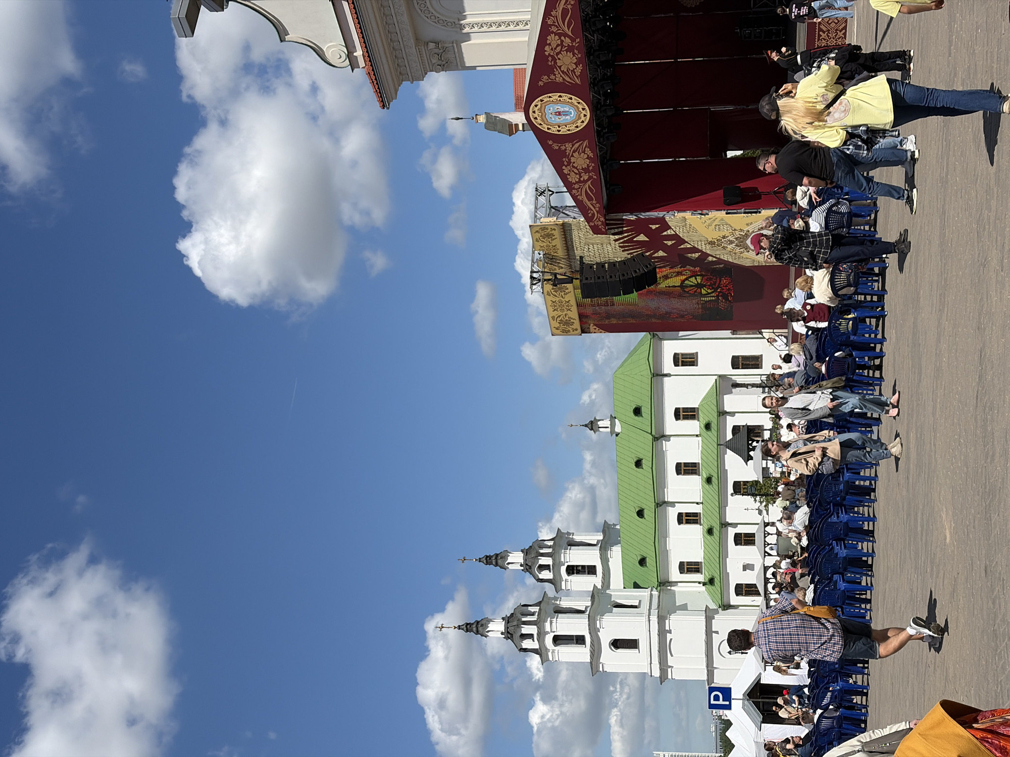 The image shows a scene in Minsk, Belarus. The Holy Spirit Cathedral is visible, along with a stage set up in front of it. People are seated in rows of blue chairs, facing the stage. Some individuals are standing near the stage. The sky is blue with fluffy white clouds. A parking sign is visible.